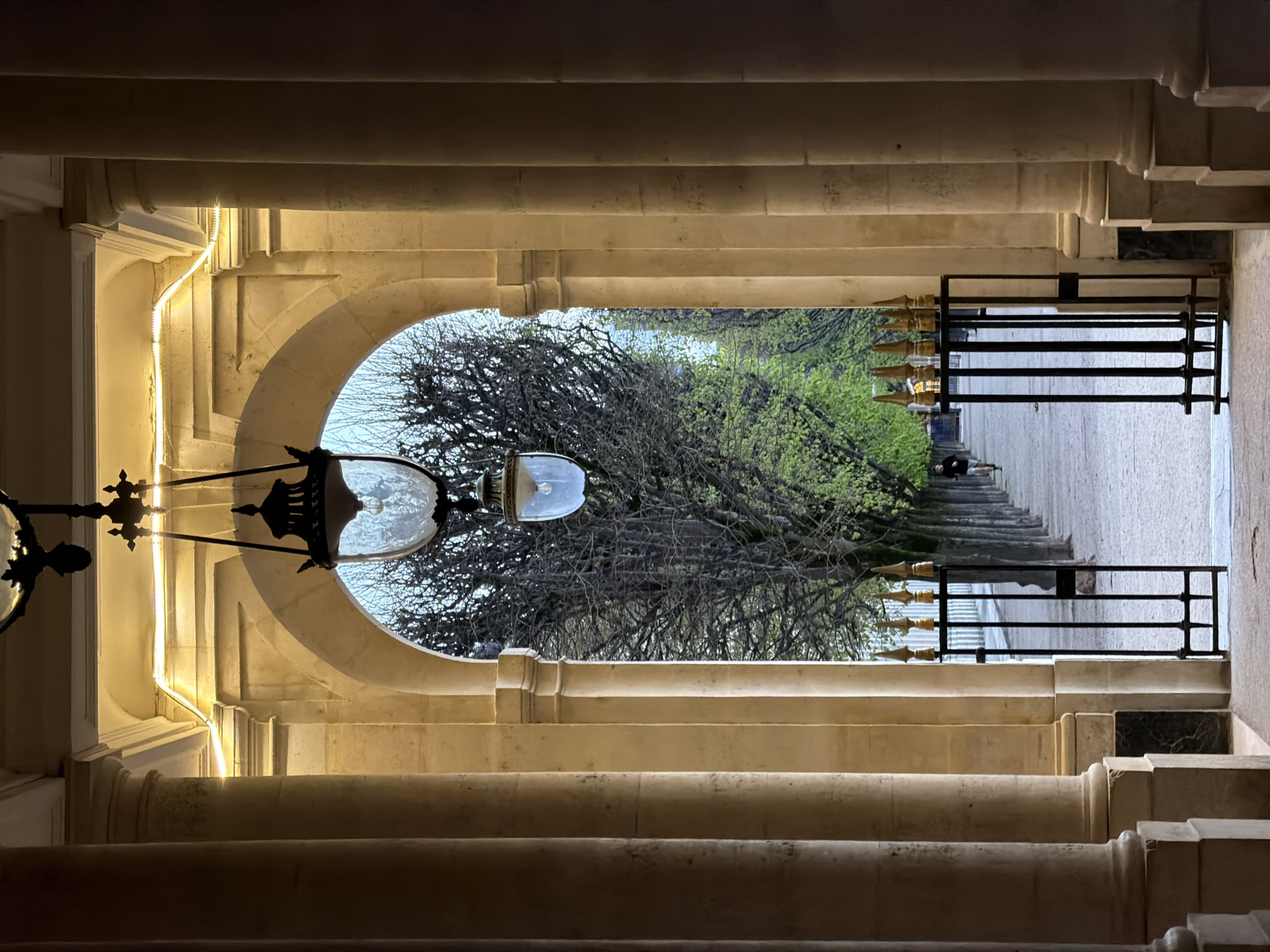 The image depicts a view looking through a stone archway into the Jardin du Palais Royal in Paris, France. The foreground features a vaulted stone arcade, supported by large, light-colored classical columns with fluting. A strip of warm, yellow LED lighting is installed along the inner curve of the archway. Two ornate, dark metal lanterns with textured glass globes and visible incandescent bulbs hang centrally within the arch, illuminating the space. At the threshold, a black metal gate, partially open, stands with vertical bars and golden spire finials. Beyond the gate, a wide, light-colored gravel path extends into the distance, bordered on both sides by rows of trees. The trees closer to the viewer have bare, dark branches, while those further down the path exhibit vibrant green spring foliage. A single person, dressed in dark clothing and light shorts, is visible running away from the viewer along the central path. The background shows an overcast or twilight sky above the distant trees.