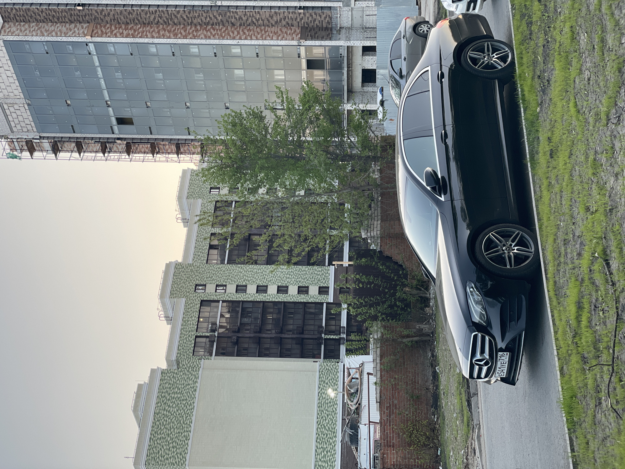 The image depicts a black Mercedes-Benz E-Class parked on a street adjacent to modern apartment buildings in Kazan, Russia. A building under construction is visible in the background. The license plate of the car is partially visible and suggests it is a Russian vehicle. The sky is overcast. The ground shows a mix of asphalt and grassy areas. Another car is parked behind the Mercedes-Benz. A brick wall and some vegetation can be seen between the Mercedes-Benz and one of the buildings. The buildings are constructed in a contemporary architectural style, with a mix of glass and textured facades.
