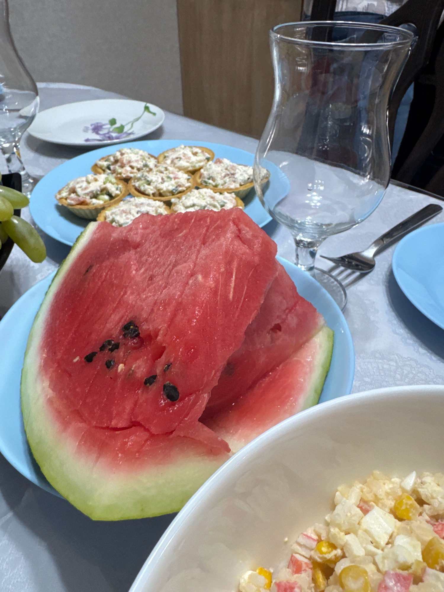 The image captures a close-up view of a table setting, likely in a home environment. The foreground is dominated by a slice of watermelon served on a light blue plate. The watermelon is vibrant red with visible black seeds and a light green rind. Adjacent to the watermelon is a white bowl filled with a salad. This salad appears to contain ingredients such as corn kernels, diced white pieces (possibly potato or cheese), and red and white shredded pieces (likely imitation crab meat).
Further back on the table, another light blue plate holds several small tartlets filled with a creamy salad mixture, possibly a seafood or egg-based salad. A white plate with a delicate floral design is visible to the left of the tartlets. A clear glass goblet, designed with a flared rim and a slender stem, stands empty to the right of the tartlets, hinting at a beverage to be served. A silver fork rests near the goblet. In the background, a light-colored wooden surface is visible, potentially a table or cabinetry. A bunch of green grapes is partially visible on the left side of the image.
The lighting is soft and even, suggesting an indoor setting, possibly during a meal or gathering. The overall impression is one of a prepared meal, with a focus on fresh fruits and appetizers. The setting does not provide any specific indicators of location within the city of Novosibirsk, Russia.