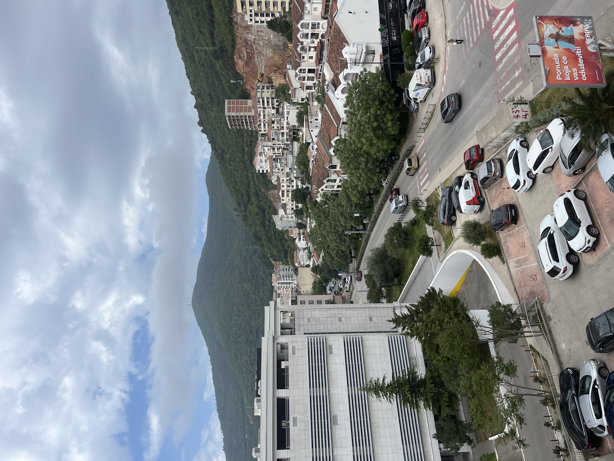 This is an elevated view of Budva, Montenegro. A large, verdant mountain rises along the left, partially obscured by cloudy sky. A densely packed section of white buildings with orange tiled roofs stretches along the mountain's base. A road lined with trees and parked cars runs parallel to the buildings. The street has a crosswalk and a large sign. A modern white building with vertical stripes occupies the foreground, along with trees and a parking area. A few people are visible on the street.