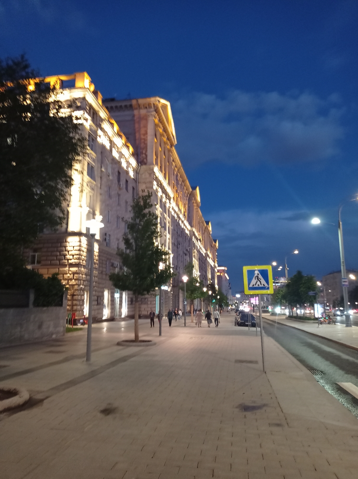 The image depicts a street scene in Moscow, Russia, at dusk. A large, ornate building with illuminated windows and architectural details lines the left side of the street. The building appears to be a Stalinist Empire style structure, common in Moscow. A sidewalk paved with interlocking stones runs parallel to the building. Trees are planted along the sidewalk. Several people are walking on the sidewalk, appearing to be pedestrians. A pedestrian crossing sign is visible on the right side of the street, indicating a crosswalk. Cars are driving on the road, and streetlights illuminate the area. The sky is a deep blue, with some clouds, suggesting it is twilight. The overall atmosphere is one of an urban evening.