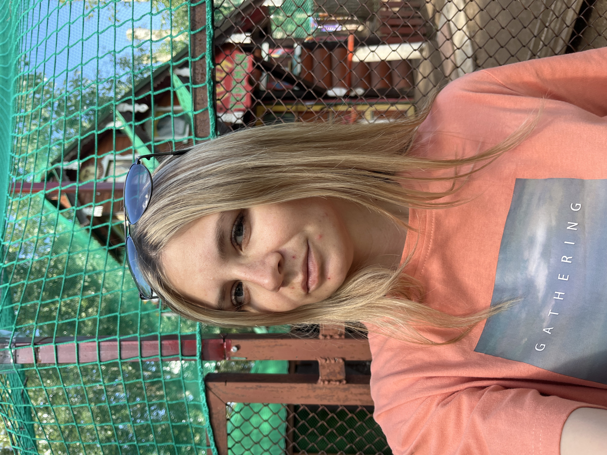 A young woman with blonde hair and sunglasses on her head is taking a selfie outdoors. She is wearing a peach-colored t-shirt with a picture and the word "GATHERING" printed on it. She has some red blemishes on her face. Behind her, there is a wooden structure with red and white trim, a green net fence, and a chain-link fence. The environment suggests a park or an outdoor recreational area. The background and the architectural style of the wooden structure indicate it is likely in Chaikovsky, Russia, a city known for its recreational facilities and Soviet-era architecture.