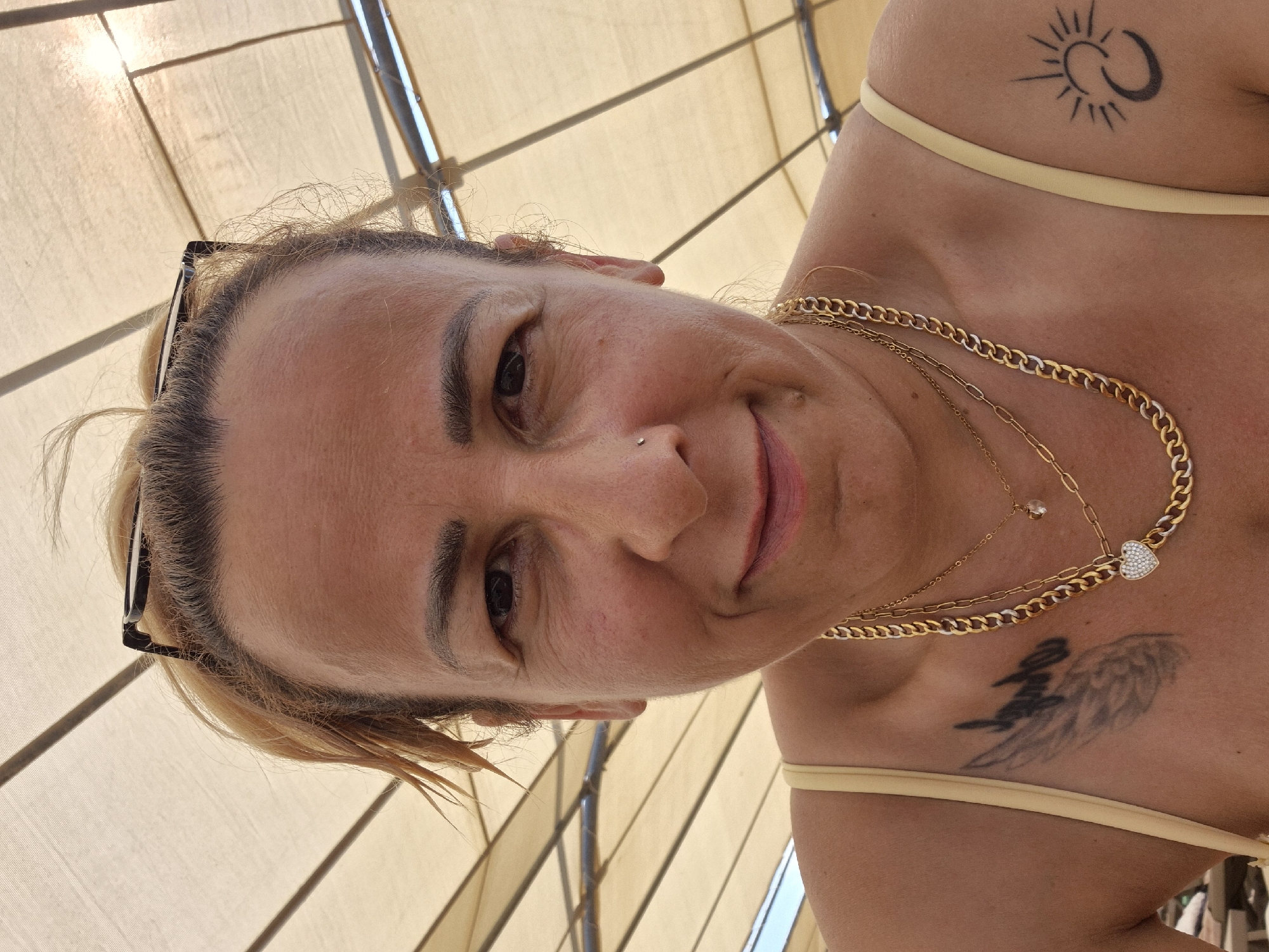 The media file shows a close-up, rotated portrait of a woman. She has blonde hair and is wearing a light yellow top, likely a bikini top, with gold and silver necklaces. One necklace features a small heart-shaped pendant. She also has a tattoo of a sun and moon on her upper left arm and a wing tattoo on her chest. She is wearing glasses on top of her head. Her face is turned towards the camera, and she has a slight smile. The background appears to be a light-colored canopy with structural supports, suggesting an outdoor setting, possibly a beach or poolside. Sunlight filters through the canopy, creating a warm, bright atmosphere. The time of day appears to be daytime, likely sunny. No text is visible in the image. The overall mood conveyed is relaxed and summery.