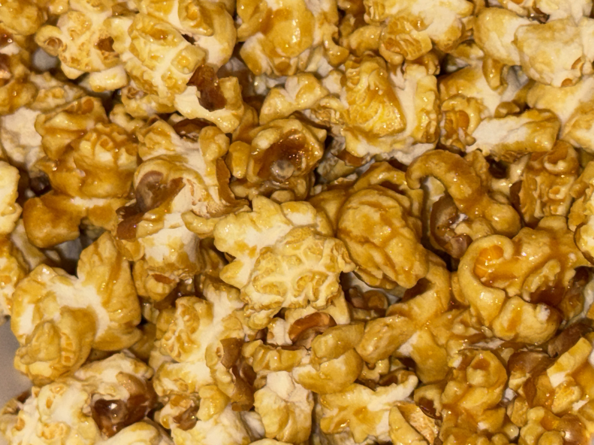 This is a close-up, high-angle shot of caramel popcorn. The popcorn is golden brown and appears to be coated in a shiny caramel glaze, with some pieces clumping together. The texture is rough and irregular, characteristic of popped corn. Small, darker brown pieces, possibly nuts or burnt caramel bits, are scattered throughout the popcorn. The lighting suggests it's an indoor setting, possibly a kitchen or a food establishment, as the caramel has a glistening sheen, implying a controlled environment. There is no visible text. The image is an unspecific, close-up view of a popular snack, evoking a sense of indulgence and comfort food. Given the location context of Dubai, UAE, this could be artisanal caramel popcorn from a local confectionery or a common snack enjoyed in the region. The time of day and weather are not discernible from the image. No people are present.