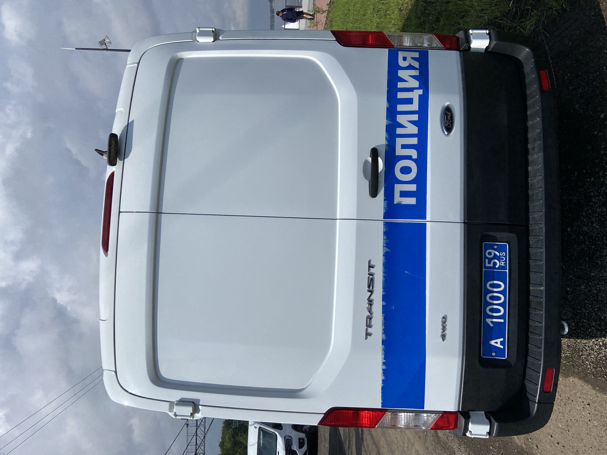 The image shows the rear of a white Ford Transit van, presumably a police vehicle, in Perm, Russia. The van has "ПОЛИЦИЯ" (Police) written in large white letters on a blue stripe across its rear doors. The license plate is "A 1000 59 RUS". A person is walking on a path in the background to the right, and another white van is partially visible to the left. The sky is cloudy.