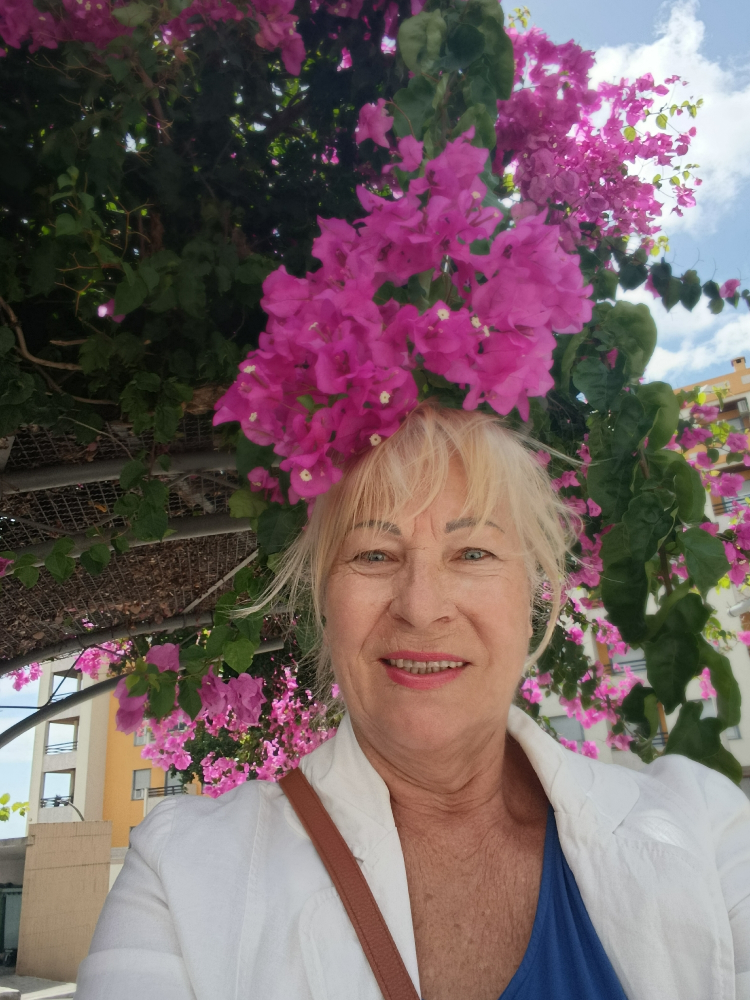 A blonde woman with blue eyes and a bright pink lipstick smiles at the camera. She is wearing a white jacket over a blue shirt and a brown strap is visible over her right shoulder. She is positioned in front of a vibrant bougainvillea plant with pink flowers. In the background, there are buildings with balconies and a cloudy blue sky. The woman's head is framed by the bougainvillea flowers. The scene suggests an outdoor setting in a warm climate, possibly a city like Setúbal, Portugal, given the architectural style of the buildings.