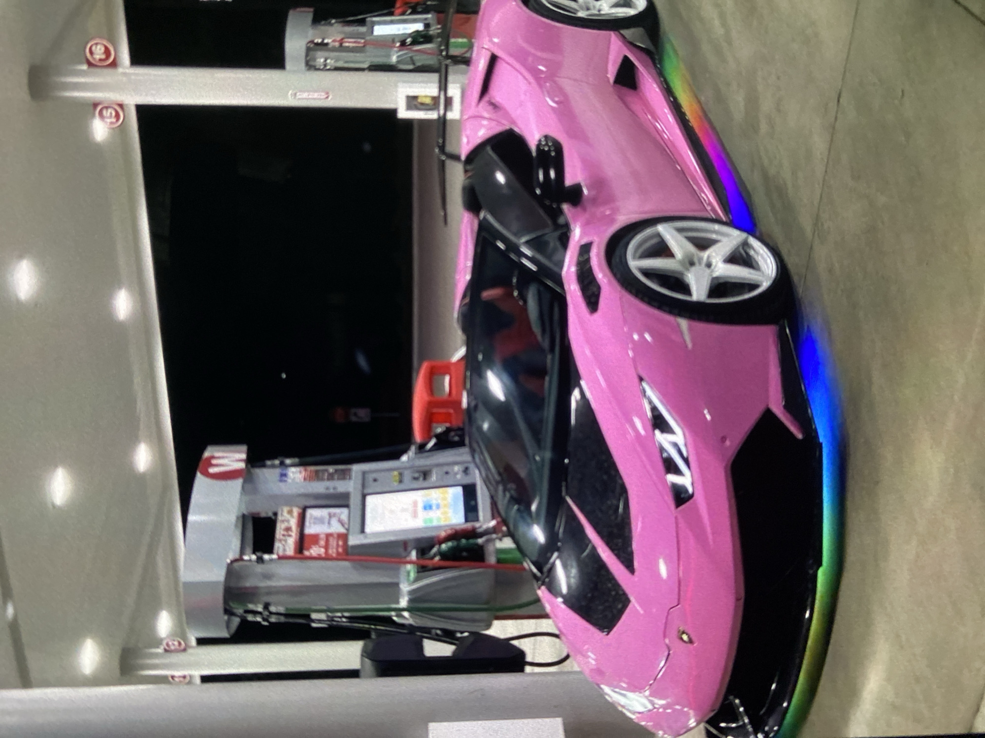 The image shows a pink Lamborghini parked at a gas station. The gas station has a gray and white awning, and the gas pumps are visible with their screens displaying numbers. The car has rainbow-colored edging and custom rims. The background is dark, suggesting nighttime. There is no indication of this image's location being in the city of Lhokseumawe, Indonesia. There are no people visible in the image.