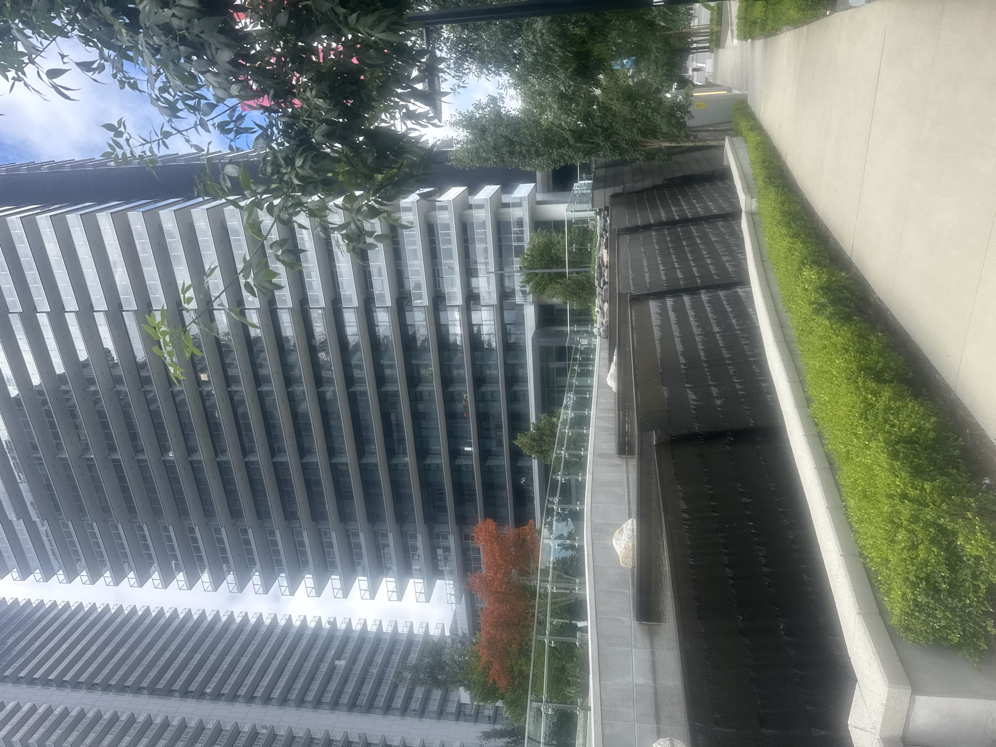 The image depicts a modern urban scene in Burnaby, Canada, likely in a residential or mixed-use development. Dominating the background are two towering, multi-story condominium buildings with repetitive balconies and glass facades, reflecting the overcast sky. In the foreground, a tiered water feature cascades down dark, textured walls, creating a tranquil atmosphere. A paved walkway runs alongside the water feature, bordered by meticulously manicured green hedges. Several trees are present, including a vibrant red-leafed tree indicating an autumnal season. The overall impression is one of contemporary urban design, integrating natural elements with high-rise architecture. There are no people or discernible actions visible in the frame.