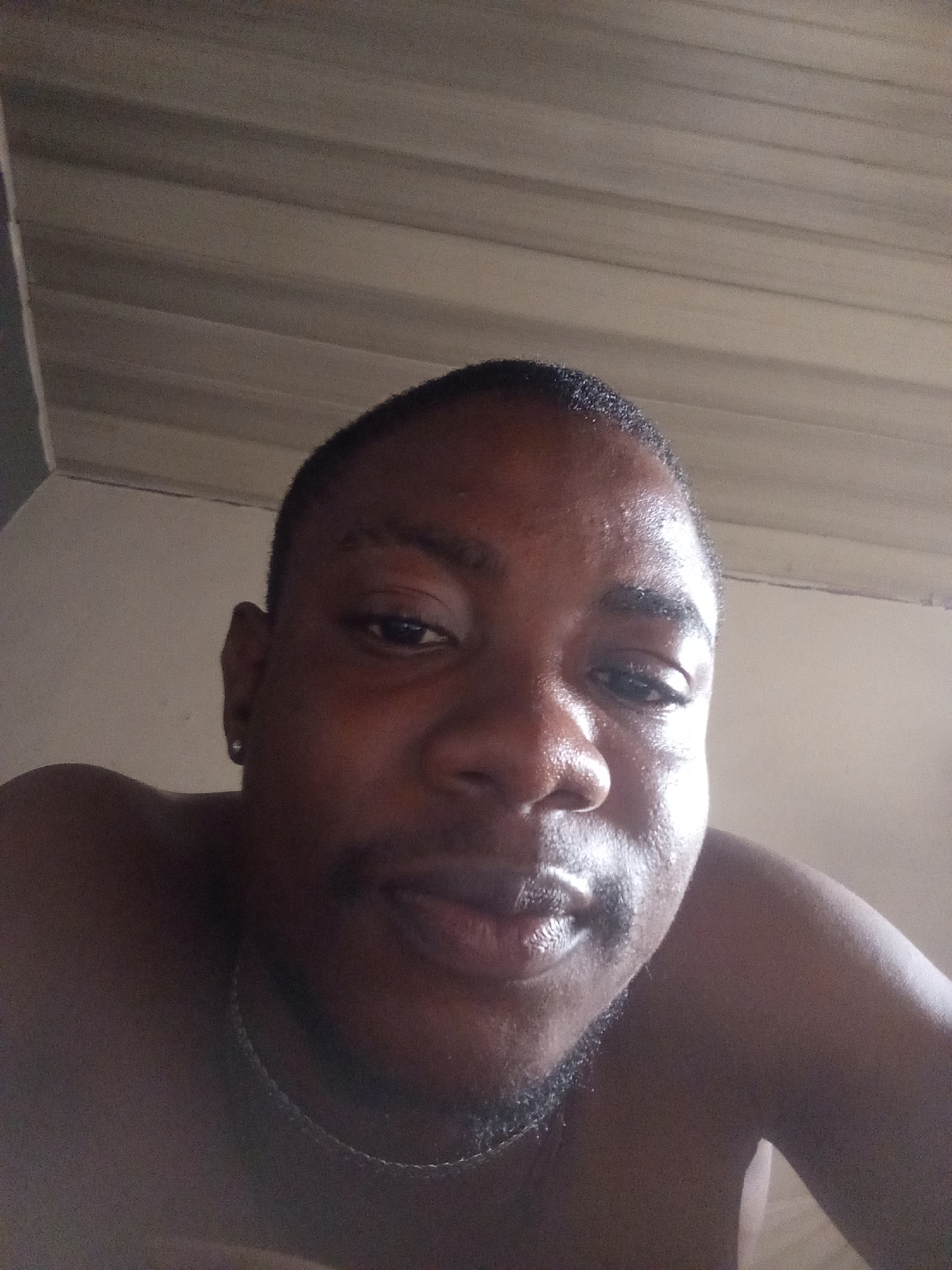 This is a close-up selfie of a young Black man, taken from a low angle. He is shirtless and appears to be indoors, with a corrugated ceiling visible in the background. The lighting is dim, with some reflections on his skin. He has a short haircut, a light beard, and is wearing a thin chain necklace. There is a small earring visible in his left ear. The image does not contain any specific details that would allow for the identification of the location within Ashata, Nigeria.