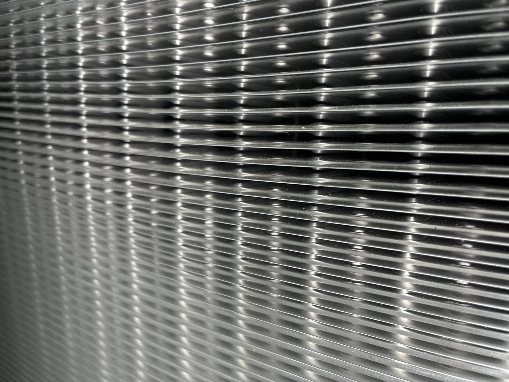 The image shows a close-up, abstract view of a metallic surface with a repeating grid pattern. The surface is made of parallel, horizontal ridges that create a series of indentations or channels. Within these channels, there are smaller, circular raised bumps that also form a regular pattern. The lighting highlights the reflective nature of the metal, creating bright glints and deep shadows that emphasize the texture and depth of the surface. The perspective is angled, causing the horizontal lines to appear to converge slightly towards the right side of the frame, giving a sense of depth. The overall impression is one of industrial or structural design, possibly a ventilation grate, a textured panel, or a component of a machine. There are no people, specific objects, or environmental cues to suggest a particular activity or scene. The time of day, weather, and location (Kyiv, Ukraine) are not discernible from the visual information provided. There is no visible text in the image.