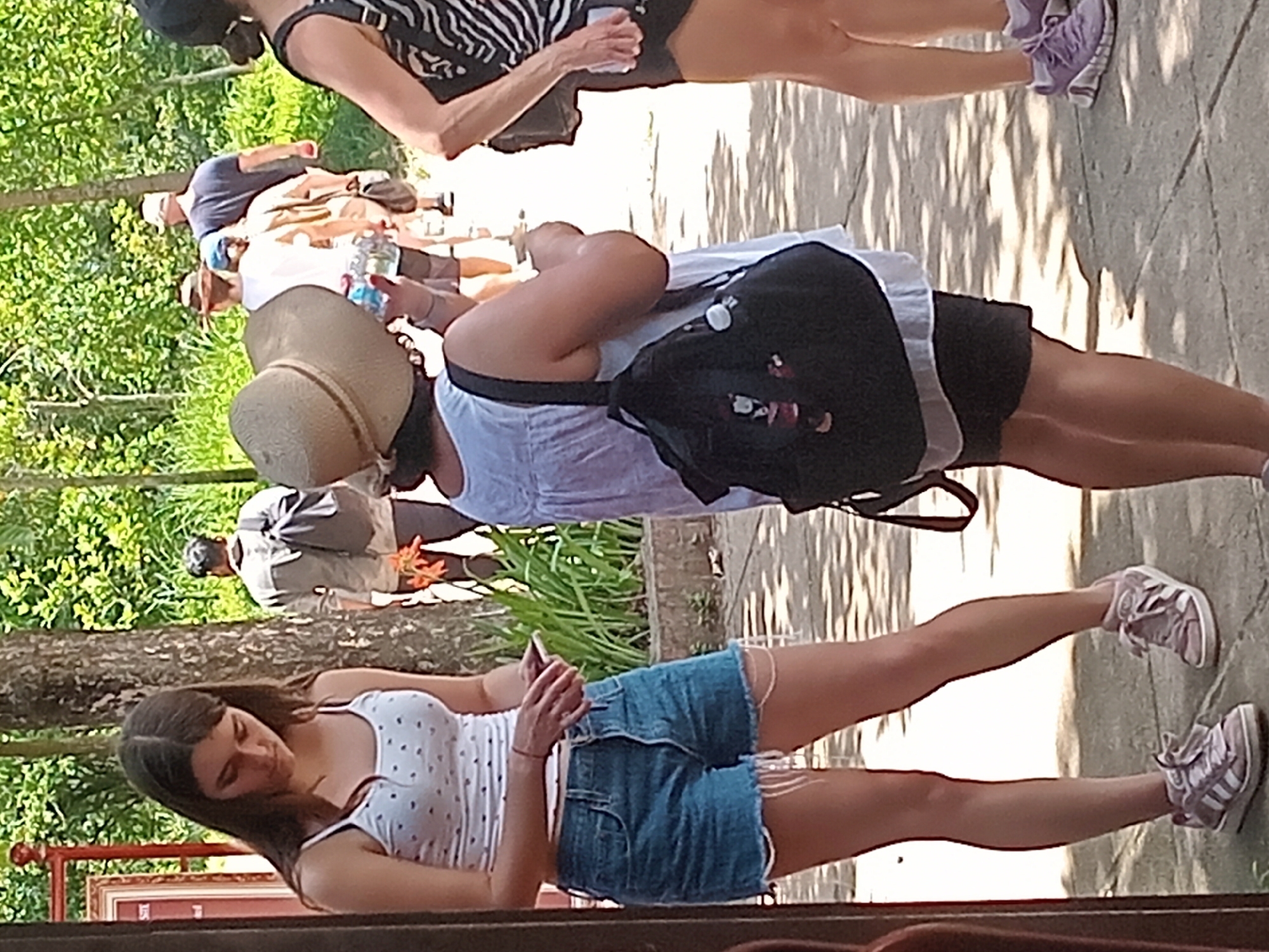 The image shows a group of people walking on a paved path outdoors. The environment appears to be a park or recreational area, with trees and greenery visible in the background. The lighting suggests it is daytime with sunlight creating dappled shadows on the ground.
In the foreground, a young woman is standing, looking at her phone. She is wearing a white tank top with a pattern of small red hearts, and denim shorts. She has on light pink athletic shoes with white laces and white stripes.
To her right, another woman is walking away, facing the right side of the frame. She is wearing a light-colored dress and a wide-brimmed straw hat. She is carrying a black backpack adorned with charms, and holding a clear plastic bottle in her hand.
Further in the background, other individuals are visible, including a man wearing a light-colored shirt and backpack, and another person with a dark blue shirt and a cap. A woman in a black and white striped top is partially visible on the far right.
The image does not contain any specific landmarks or features that would allow for precise identification of the location within Đà Nẵng, Vietnam.