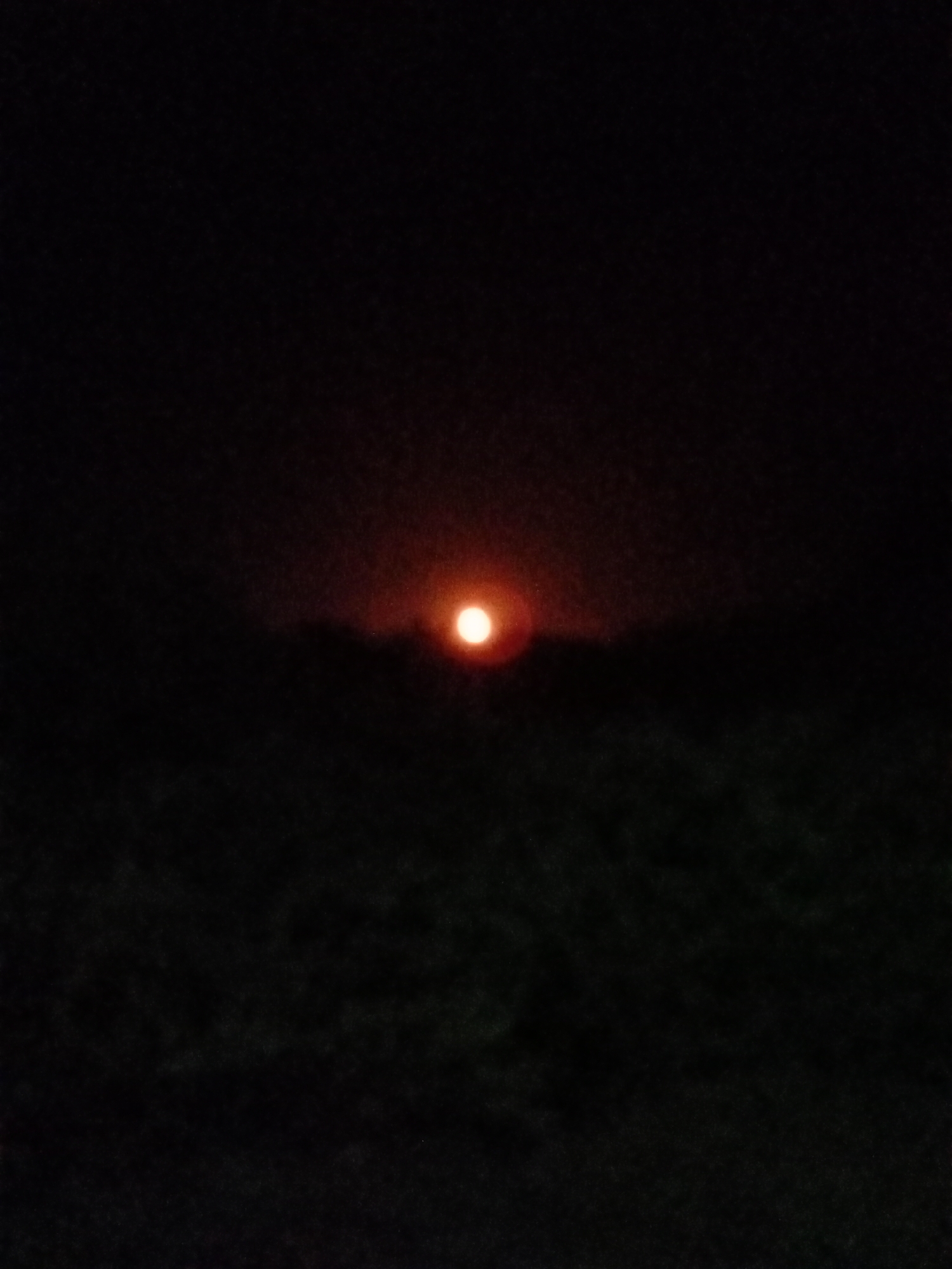 The image shows a bright orange light, likely the sun or moon, positioned on the horizon against a dark sky. The lower portion of the image appears to be a darkened landscape, possibly vegetation or terrain, with minimal discernible detail due to the low light conditions. The location of Lhokseumawe, Indonesia, is not visually confirmed in the image.