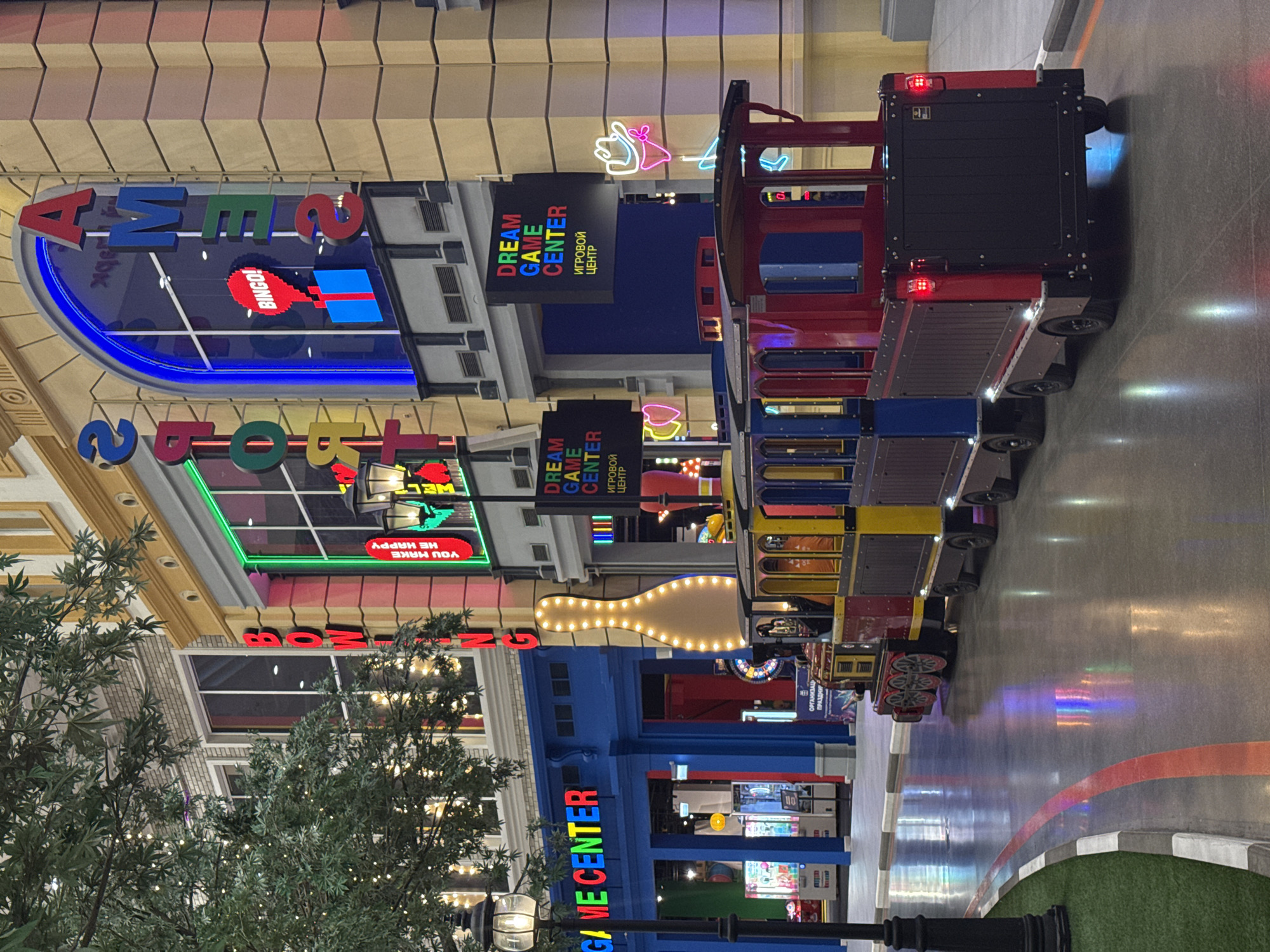 Here is a detailed description of the image:
**Scene:** The image shows the interior of what appears to be an indoor entertainment center or amusement park setting. There are several brightly colored signs with words that read "GAME CENTER" in various designs and configurations. The signs are illuminated with neon lights and are placed on building facades. There is a train consisting of multiple cars with colorful designs (red, blue, yellow), stopped on what looks like a painted road inside the entertainment center.
**Objects:** Besides the signs and the train, there is a bowling pin, also illuminated with lights, serving as a decorative element. A faux tree is in the foreground, adding to the illusion of an outdoor setting. The path is painted with red and orange colors and has green patches with white and black borders.
**Environment:** The overall environment is bright and colorful, simulating an outdoor urban scene but contained within an indoor space. The image is rotated counter-clockwise.