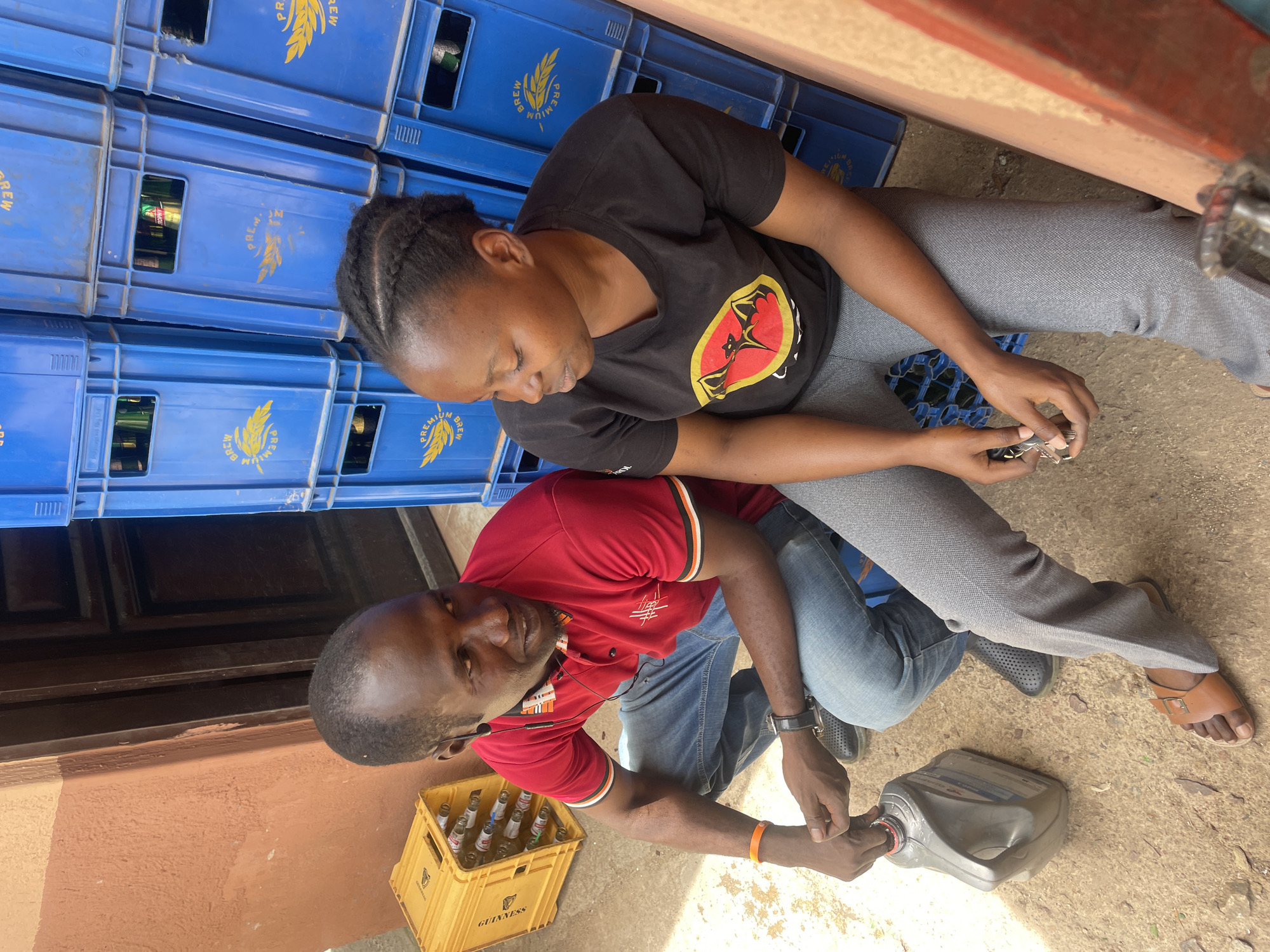 The image captures two individuals, a woman and a man, engaged in an activity outdoors during daylight.
The woman is seated on the ground, leaning against a stack of blue plastic crates. She wears a black t-shirt featuring a prominent red and yellow logo resembling the Ugandan flag's crested crane emblem, paired with grey pants and black sandals. Her braided hair is visible. She looks down at her hands, where she holds what appear to be keys or small metallic objects, along with a small blue, gridded item in her lap.
The man is crouching or kneeling in front of the woman. He wears a red polo shirt with black and white striped sleeves, blue jeans, and dark closed-toe shoes. He has a watch on his left wrist and an orange band on his right. He is holding a grey plastic container, possibly preparing to open or pour from it. He looks towards the camera with a slight smile.
In the background, multiple stacks of blue plastic crates are visible, some appearing to contain bottles. The crates bear the text "PREMIUM BREW" along with a wheat stalk logo. To the man's left, a yellow plastic crate labeled "GUINNESS" holds several empty glass bottles. The ground is a mix of dirt and concrete, with some small debris. A light brown wall is visible on the left side of the frame, and a partial structure suggests a shaded outdoor area.
The scene appears to be a casual, possibly commercial or domestic setting, such as a shop entrance or backyard storage area, under bright, sunny conditions. Both individuals seem calm and focused on their actions.