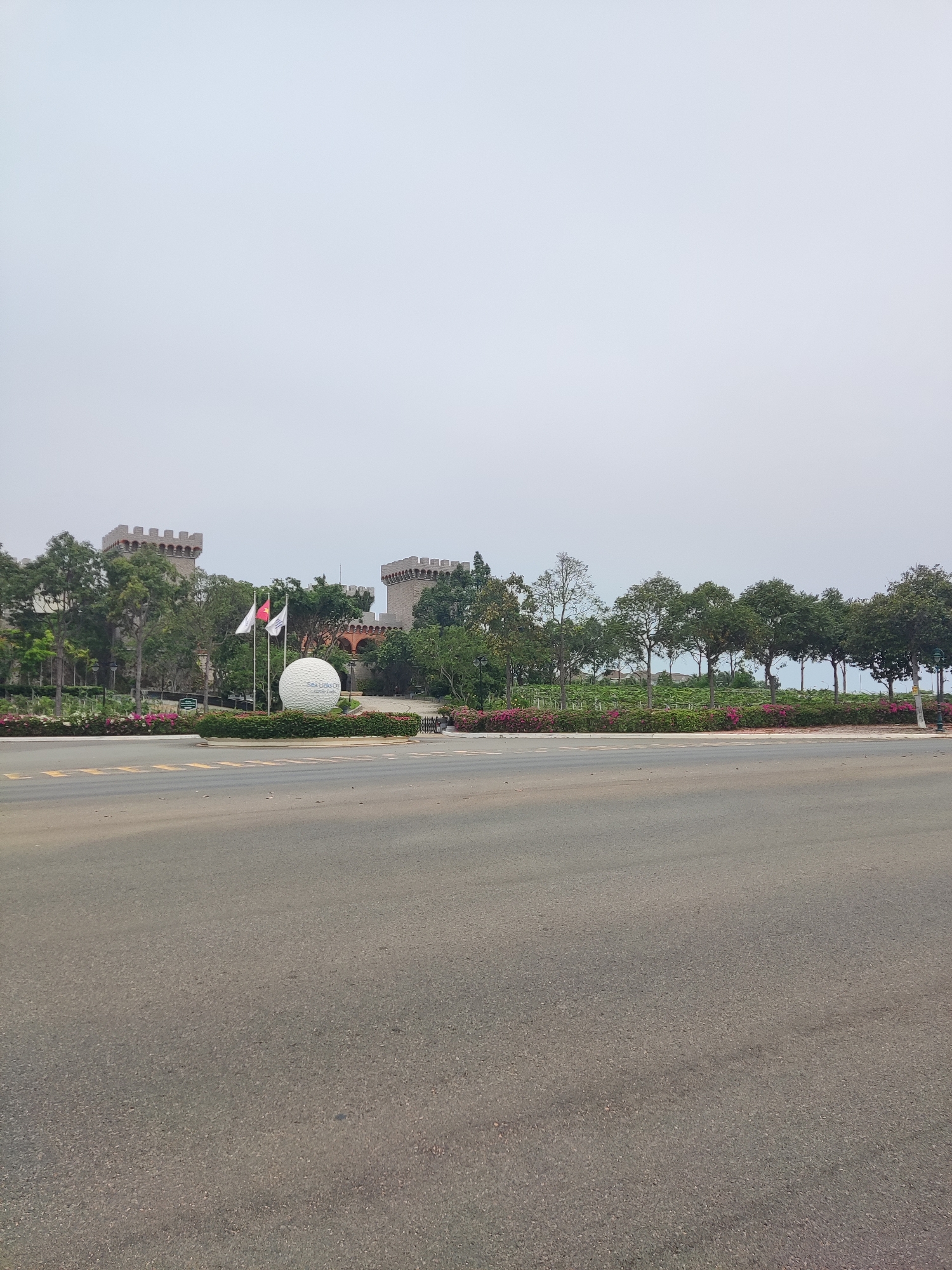 The image depicts a daytime scene in an unidentified city. A paved road occupies the foreground, stretching towards a landscaped area in the midground. Beyond the landscaping, which features trees and flowerbeds, lies a building designed to resemble a castle, with two prominent towers at either end. In front of the castle, there is a large, white spherical object. Two flags are visible near the sphere. The sky is overcast, contributing to the overall muted lighting of the scene. There are no visible people or vehicles in the image.