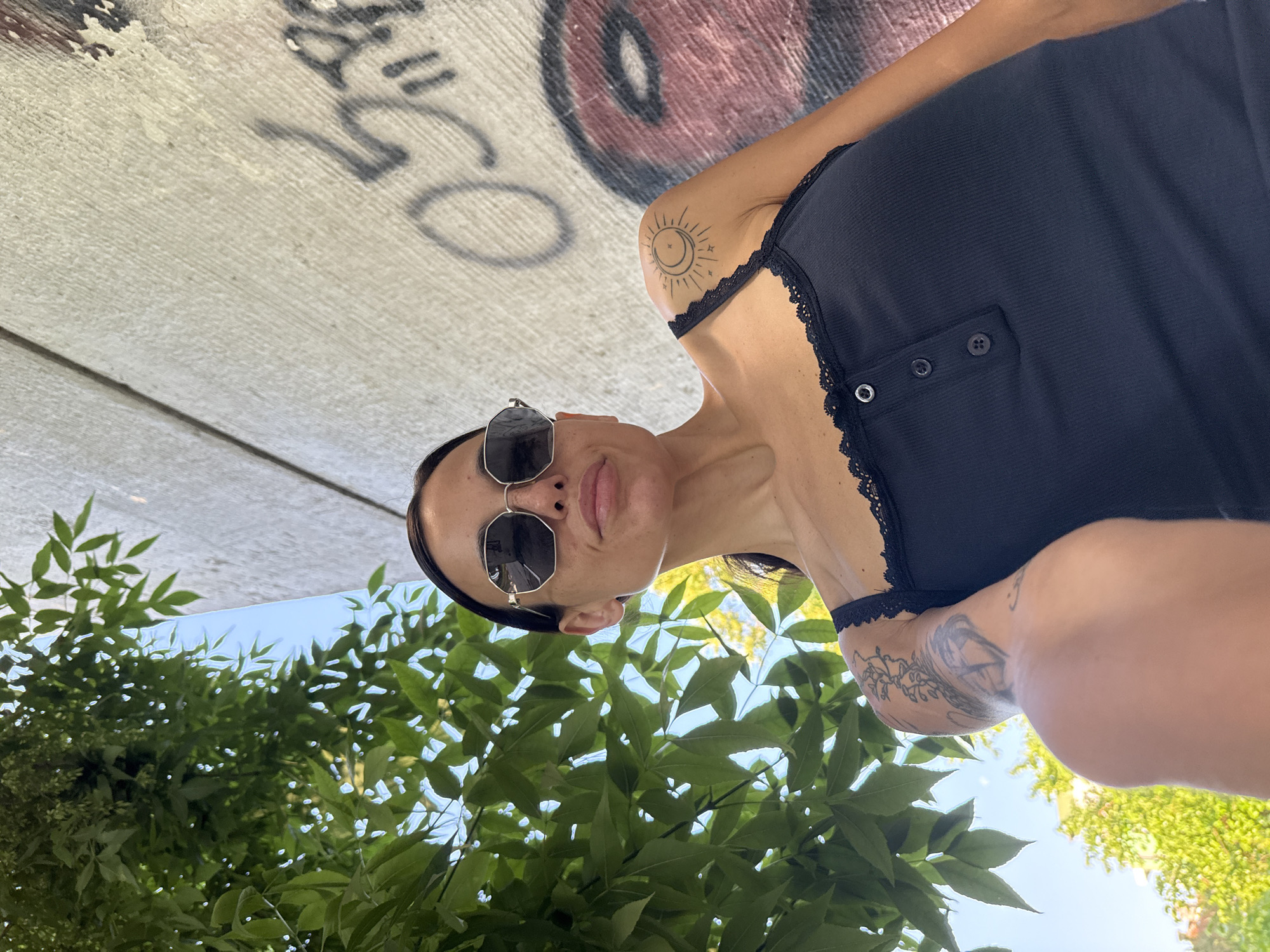 A young woman is captured in a low-angle, close-up selfie, oriented as if looking upwards towards the camera. She has dark hair pulled back and wears dark hexagonal sunglasses. Her lips are slightly parted in a neutral expression. She is wearing a black, ribbed tank top or camisole with delicate black lace trim along the neckline and straps.
Notable tattoos are visible on both arms. On her left shoulder, there is a design featuring a stylized sun enclosing a crescent moon, surrounded by radiating lines and stars. On her right forearm, a more intricate, organic or floral pattern is visible.
The background above her (in the corrected orientation) consists of a rough concrete surface, likely the underside of a bridge or overpass, heavily adorned with graffiti in black and red spray paint. Large, abstract letterforms and a prominent red circle are discernable. Below and around her are lush, vibrant green leaves of trees or bushes, with patches of bright blue sky visible through the foliage, indicating a sunny outdoor setting during daytime. The overall composition suggests an urban environment.