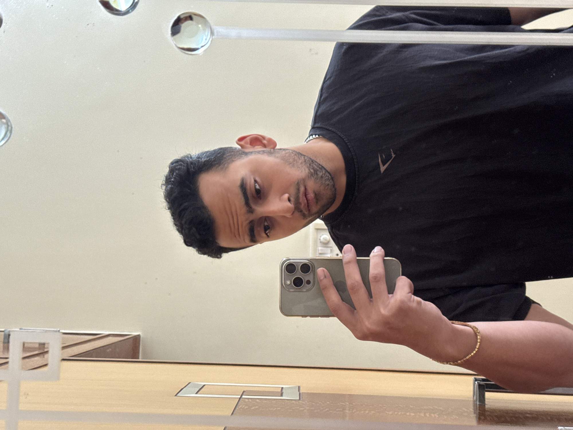 A man is taking a selfie in a mirror. He is wearing a black t-shirt and a gold bracelet. He is holding a phone with three cameras. The background is a plain wall with a lighting fixture featuring spherical lights. The reflection suggests the presence of wooden surfaces and possibly other furniture. There is no evidence to determine this is Kolkata, India.