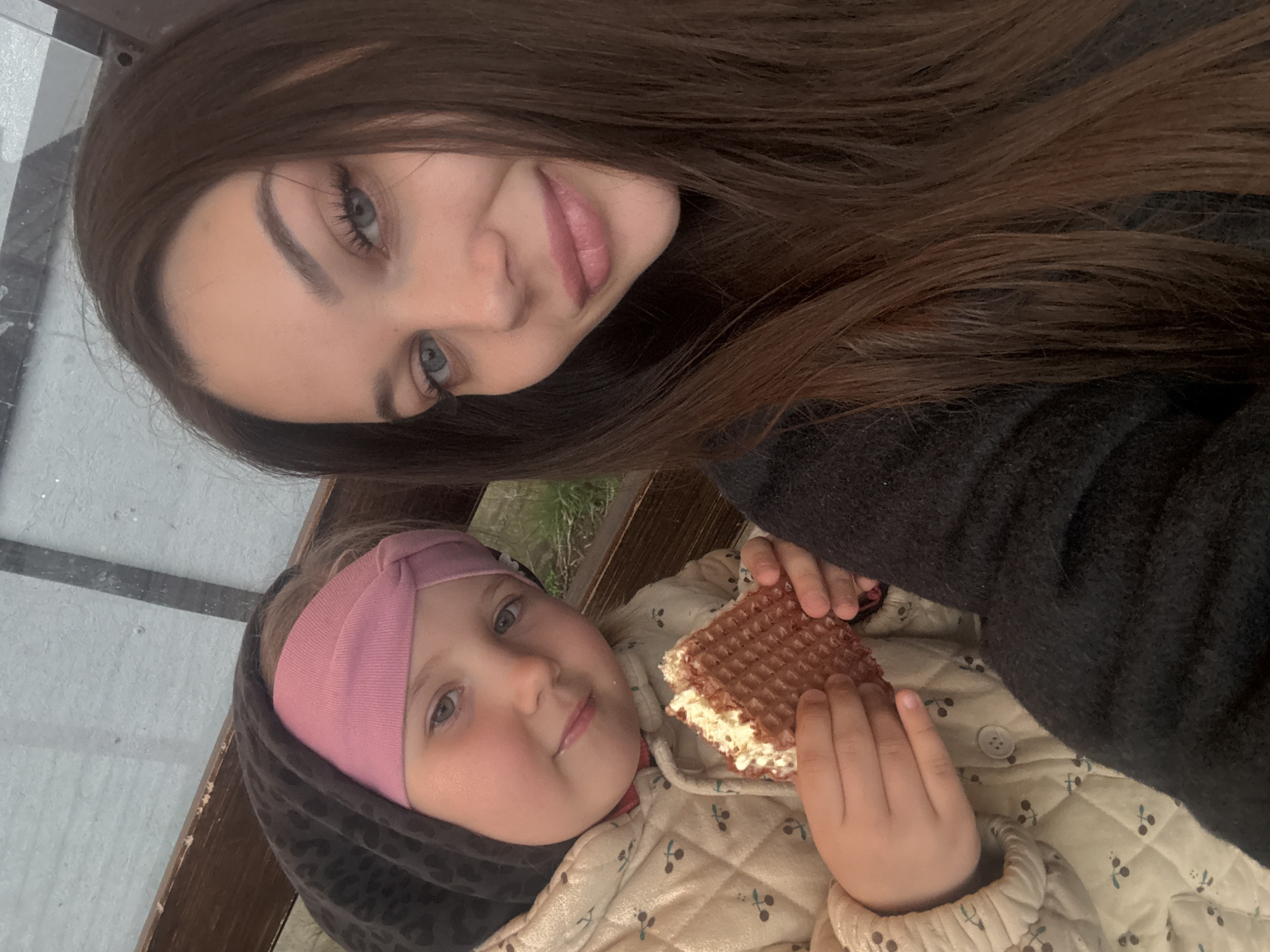 A woman and a young child are in an outdoor setting, possibly in Прикамский, Russia. The woman has long, dark brown hair and is wearing a dark-colored sweater. She is positioned at the top of the frame, looking down. The young child is lying down, wearing a pink headband and a quilted jacket with a cherry pattern. The child is holding a waffle sandwich in their hands. The background includes a window with a white frame and some greenery.