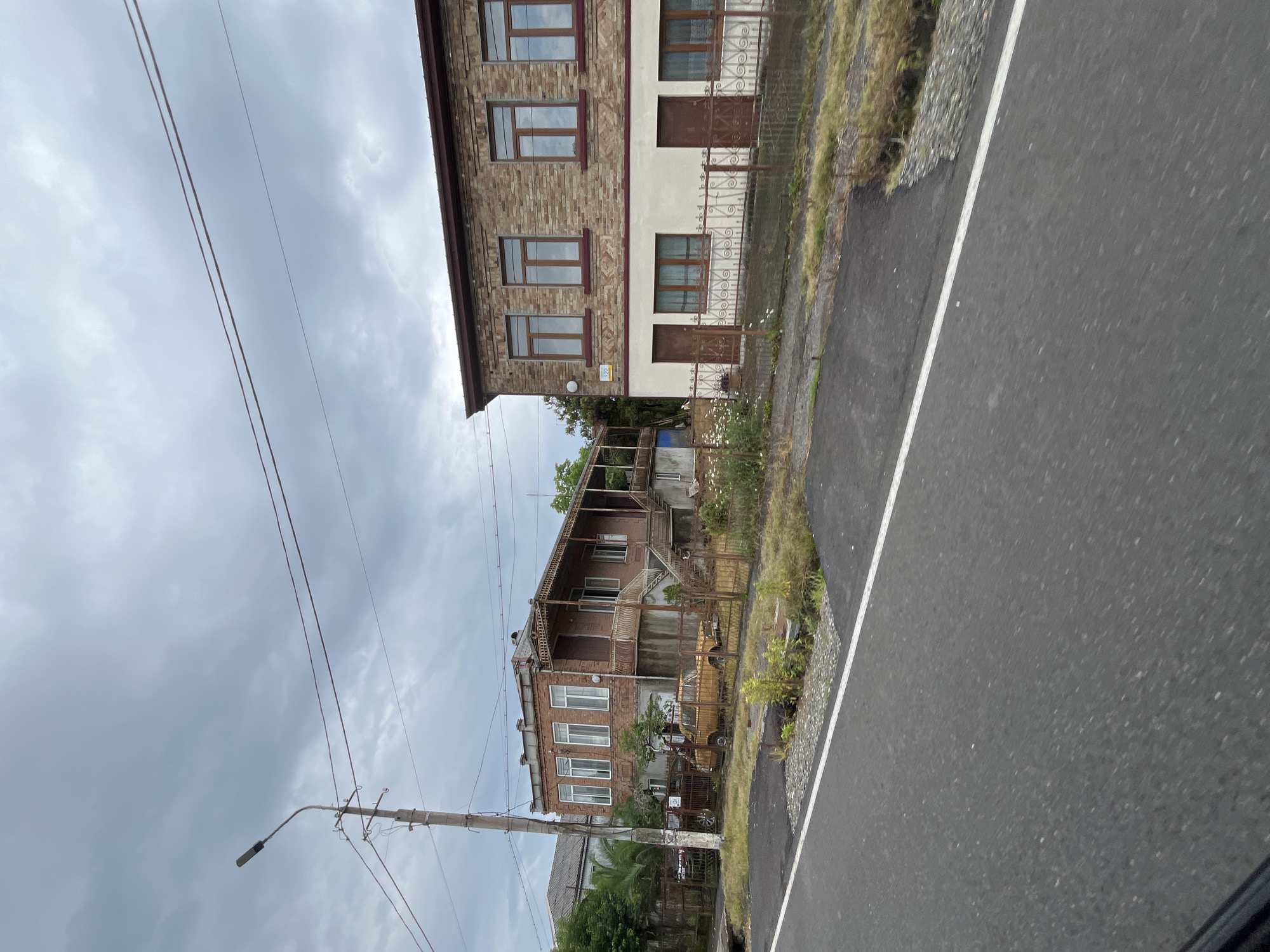 The image captures a street scene in Poti, Georgia, under a cloudy sky. The foreground features a paved road with a white line marking its edge. Adjacent to the road is a patch of gravel and then a grassy area. Behind this, a rusty metal fence separates the street from two houses.
The house on the left is a two-story building constructed with red brick. It has a covered balcony on the upper level with ornate metal railings. Several windows with white frames are visible on the facade. In front of this house, partially visible through the fence, is a weathered, yellow-colored sedan.
The house on the right is a two-story building with a facade made of brick on the upper half and a cream-colored lower half. It has several windows on the upper level with dark-colored frames. The lower level has two doors and three windows, also with dark frames. A decorative metal fence runs along the front of this property.
A concrete utility pole with a streetlamp and overhead wires stands near the left house. Trees and other vegetation are visible in the background, suggesting a somewhat rural or semi-urban setting. The overall impression is one of quiet residential life with older structures and a vintage vehicle adding character to the scene.