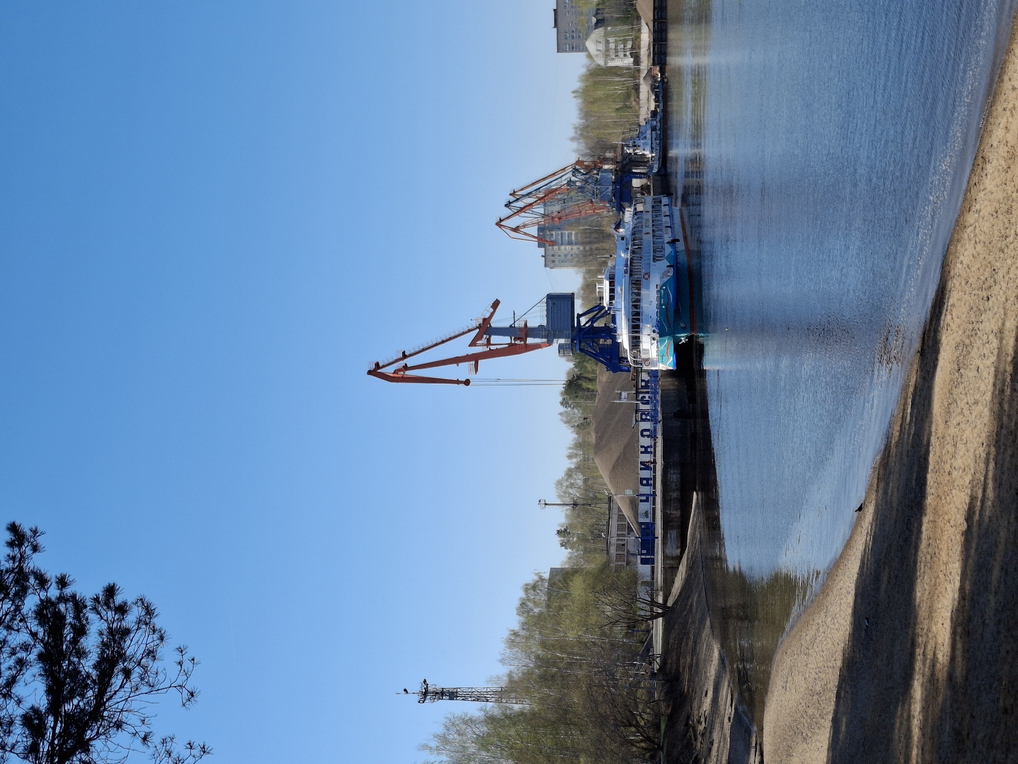 The image depicts a waterfront scene in Chaikovsky, Russia. Several large cranes are visible along the pier, indicating a port or industrial area. A passenger boat is docked alongside the pier. The water is calm, and a sandy shore is visible on the right side of the frame. In the background, there are buildings and trees lining the waterfront. The sky is clear and blue. The name "Чайковский" (Chaikovsky) is visible in Cyrillic on a structure along the pier.