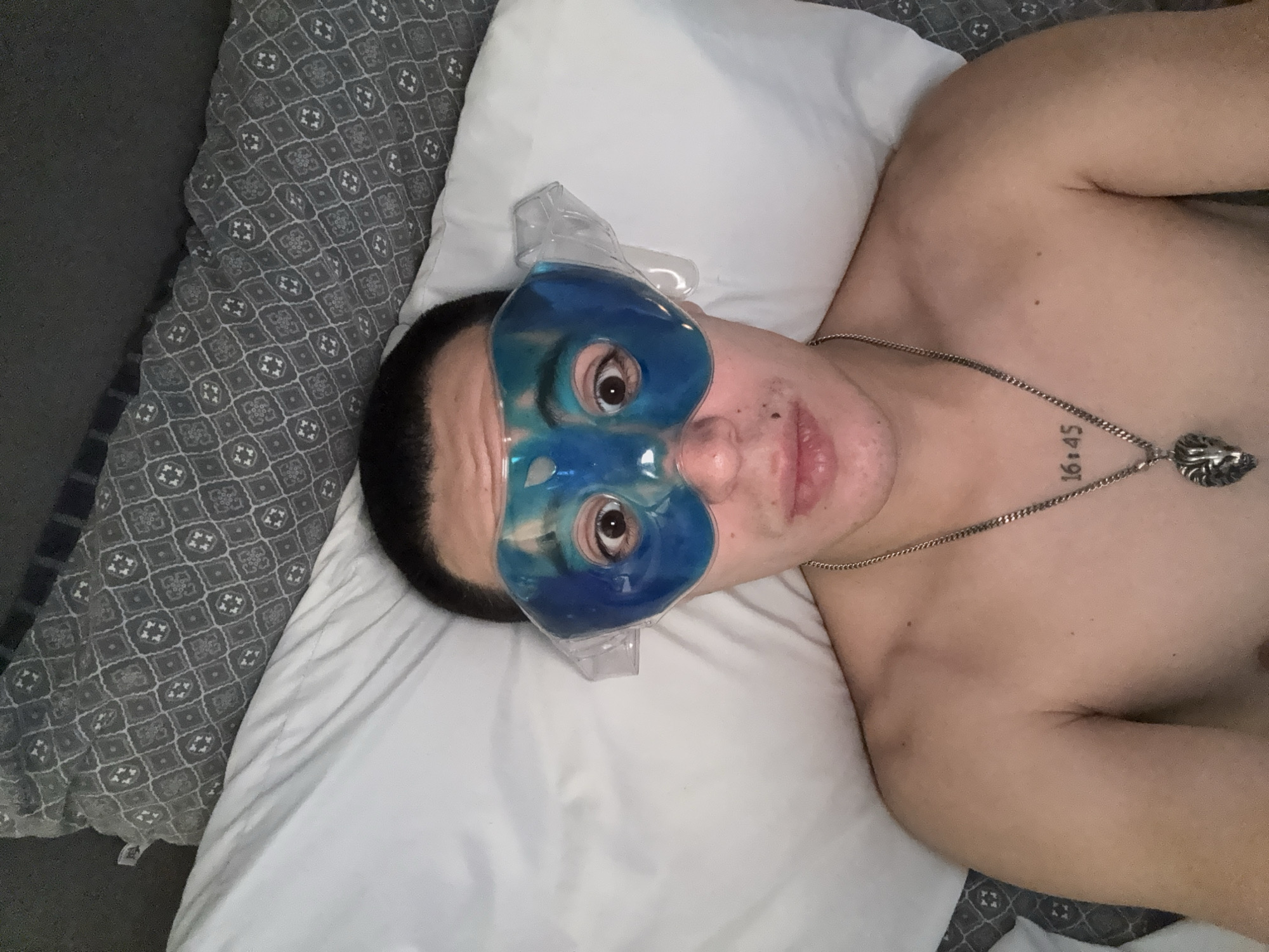 A shirtless person wearing a blue gel eye mask is lying on a white pillow on a bed with a patterned gray bedspread. The person is wearing a silver necklace with a lion head pendant. They also have a tattoo on their upper chest, visible near the necklace. The background shows part of the gray bedspread. The exact location in the city of San Bernardino Tlaxcalancingo, Mexico, cannot be determined from the visual data provided.