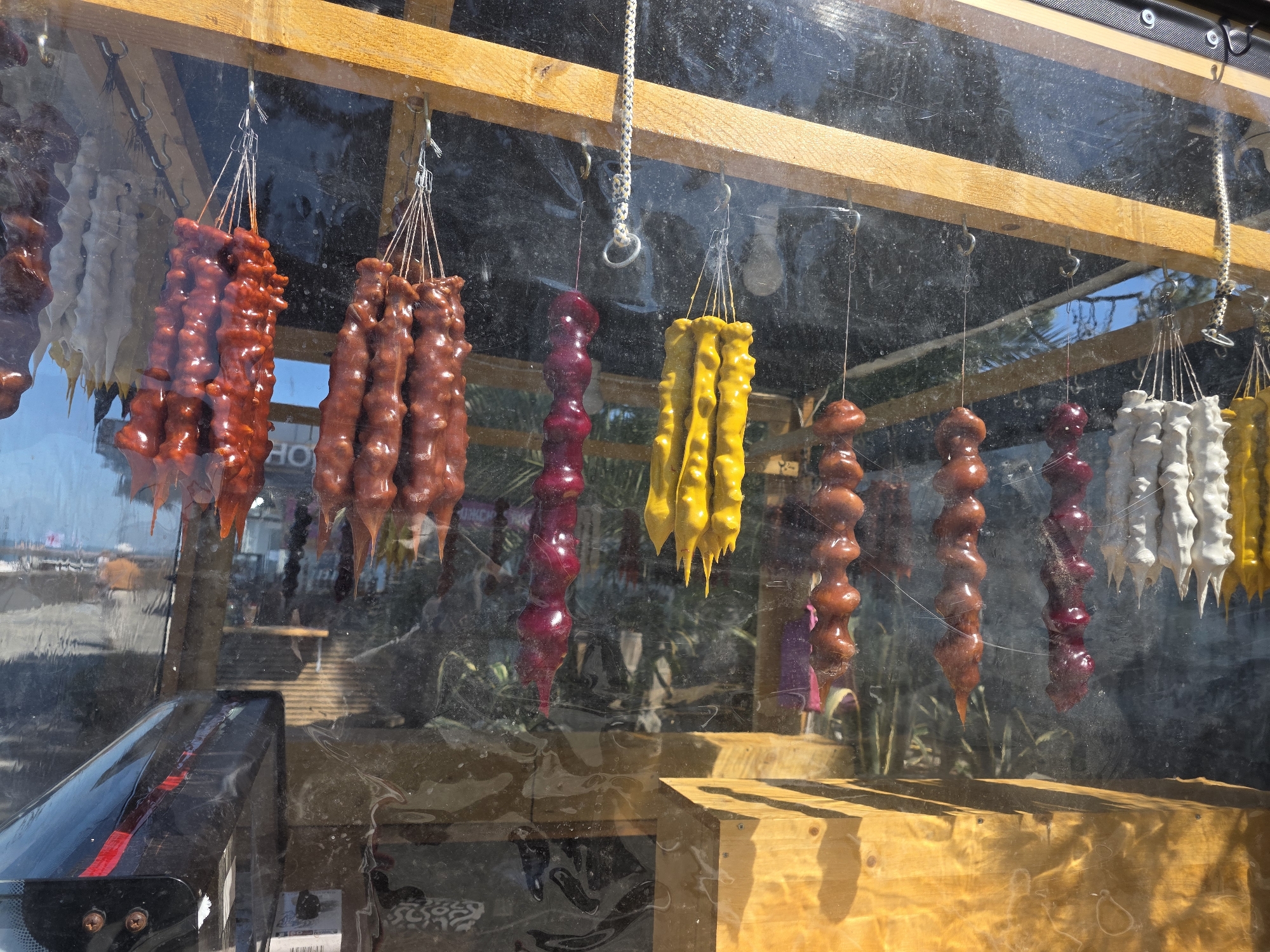 The image displays an outdoor stall or kiosk, viewed through a somewhat smudged transparent barrier, likely plastic or glass. Suspended from hooks attached to a light brown wooden beam are multiple strands of Churchkhela, a traditional candle-shaped confection.
From left to right, the visible Churchkhela strands are:
* Partial view of light grey/white strands.
* Four prominent strands of deep reddish-brown Churchkhela.
* One dark red/maroon strand.
* Two bright yellow strands.
* One medium brown strand, partially obscured.
* Further to the right, individual shorter, rounded Churchkhela-like items in medium brown and dark red are suspended.
* Finally, partial views of white/light grey and yellow strands are visible on the far right.
The Churchkhela vary in length and thickness, exhibiting a glossy, slightly uneven texture. The hooks are metallic, and the strands are tied with thin strings.
Below the suspended items, another light brown wooden structure, possibly a counter or shelf, is visible. The transparent barrier reflects a bright outdoor environment, including a clear blue sky, distant blurry green foliage, and vague structural elements. On the lower left, a reflection of a black car dashboard or interior component with a red stripe is visible, indicating the photograph was taken from inside a vehicle. No individuals are discernible in the scene. The setting suggests a market or roadside stall in a sunny outdoor location, consistent with tourist areas in Sochi, Russia, where such regional delicacies are commonly sold.