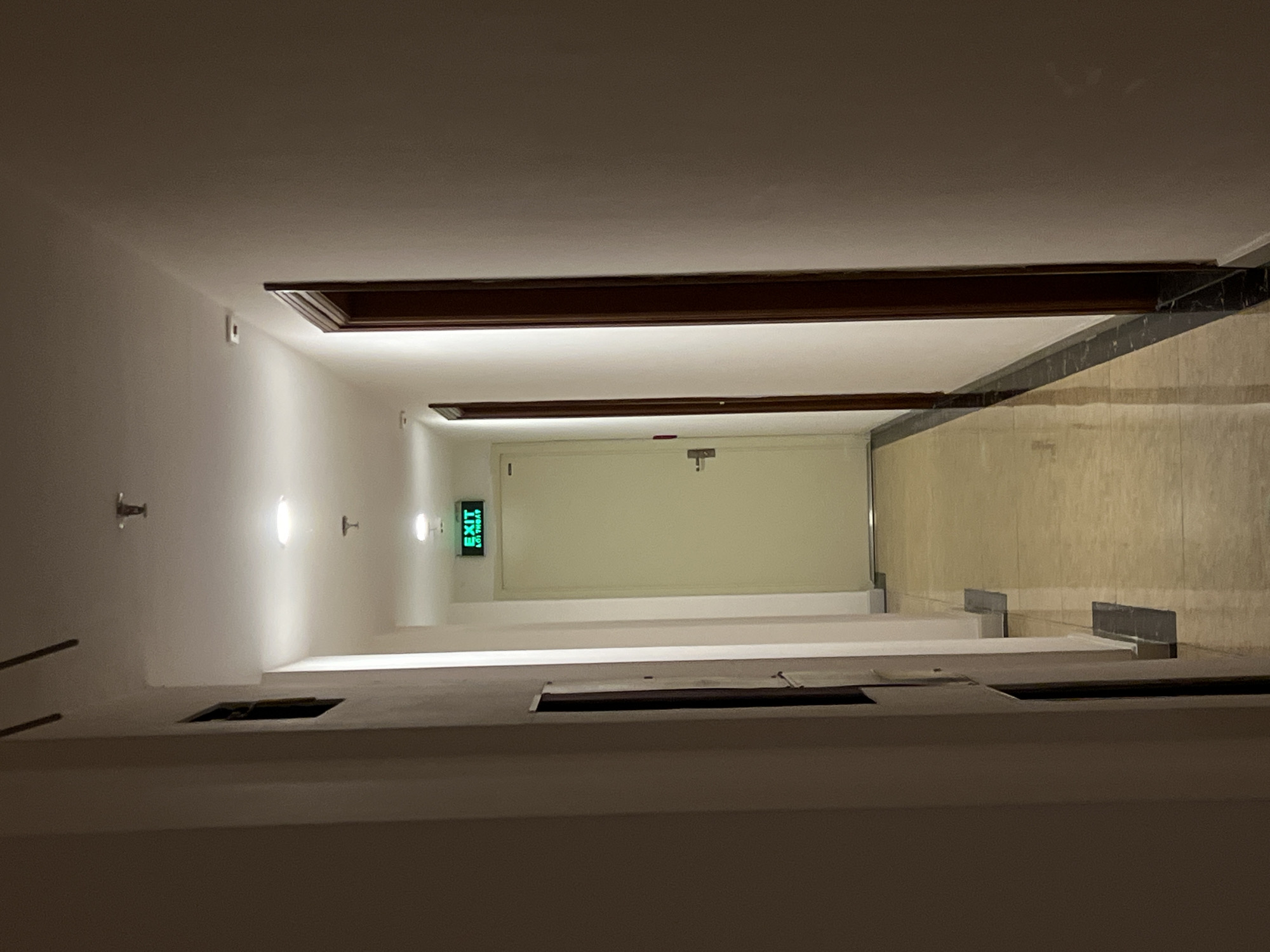 The image depicts a long, well-lit indoor hallway or corridor, viewed from a low angle looking towards its far end. At the terminus of the hallway is a closed, light-colored door.
Above and to the left of this door, a bright green "EXIT" sign is visible. The text on the sign clearly reads "EXIT" in English, and below it, "LỐI THOÁT," which is Vietnamese for 'Exit'.
The left wall features several oval-shaped, wall-mounted light fixtures casting a soft, warm glow along the corridor. Smaller, metallic, hook-like fixtures are also present on this wall. Both walls are a plain, light color, likely white or off-white. Dark brown wooden trim or decorative elements run horizontally along the top edges of the walls, creating a framed appearance further down the hallway.
The ceiling is a plain white. The floor is tiled, showing a mix of lighter and darker beige or brown sections that reflect the overhead light. On the lower right side of the hallway, a dark, rectangular opening or recessed area is visible near the floor level.
The hallway is devoid of people or any activity, conveying a sense of quietude or emptiness. The artificial lighting suggests an interior setting, likely during the evening or night, or an enclosed space. The overall scene indicates a clean, functional corridor within a building located in Bac Nha Trang Ward, Vietnam.
