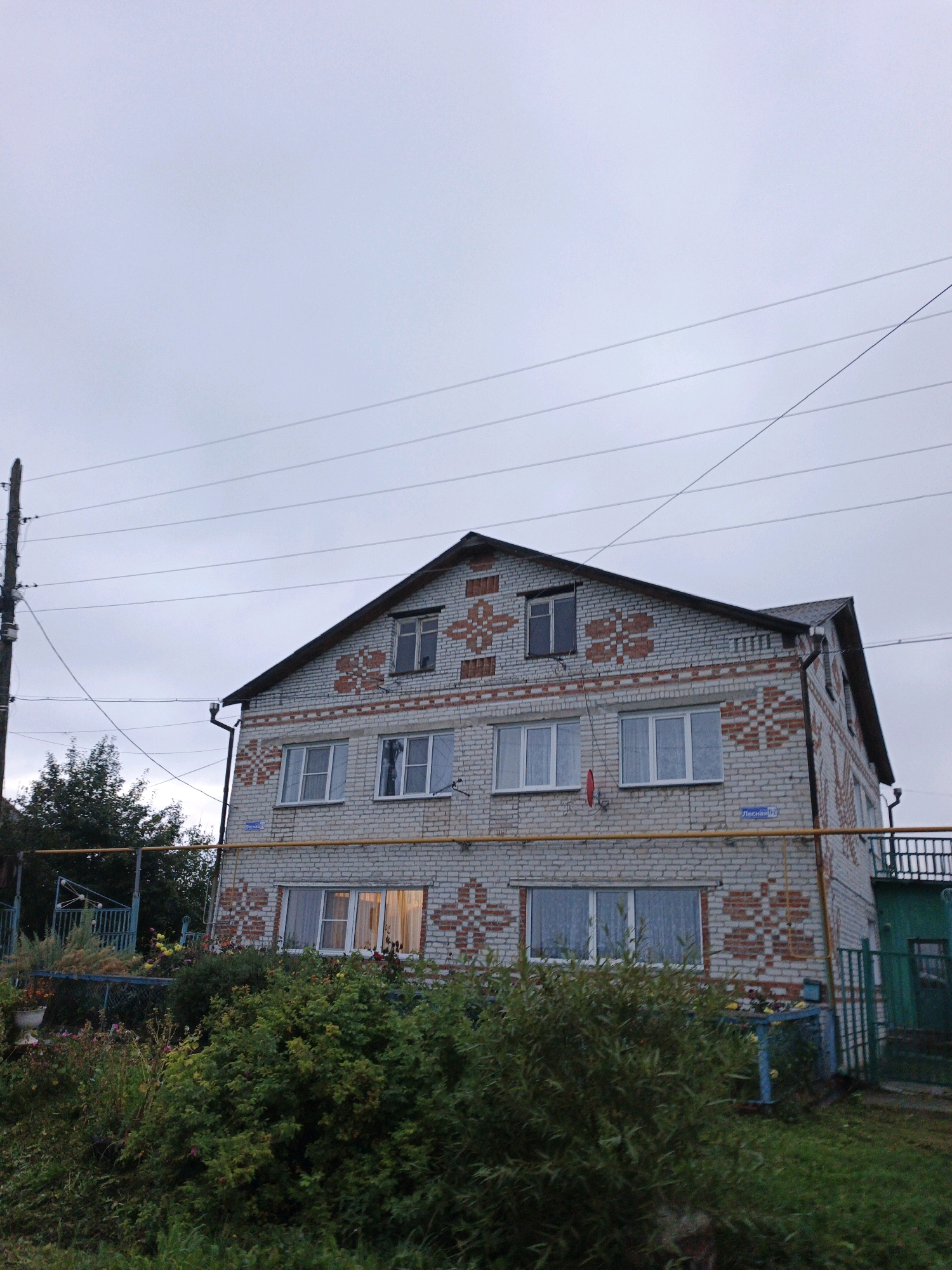 The image depicts a two-story brick building with a decorative pattern on its facade, located in Medvedyovka, Russia. The building is light grey with red brick accents forming geometric shapes on both the upper and lower levels. There are several windows visible, some with curtains. The sky is overcast and grey. In the foreground, there is lush green vegetation, including bushes and flowering plants. A wooden utility pole with multiple wires is positioned to the left of the building. A blue metal fence and a green gate are also visible. A yellow pipe runs horizontally across the facade of the building. The overall scene suggests a residential area.