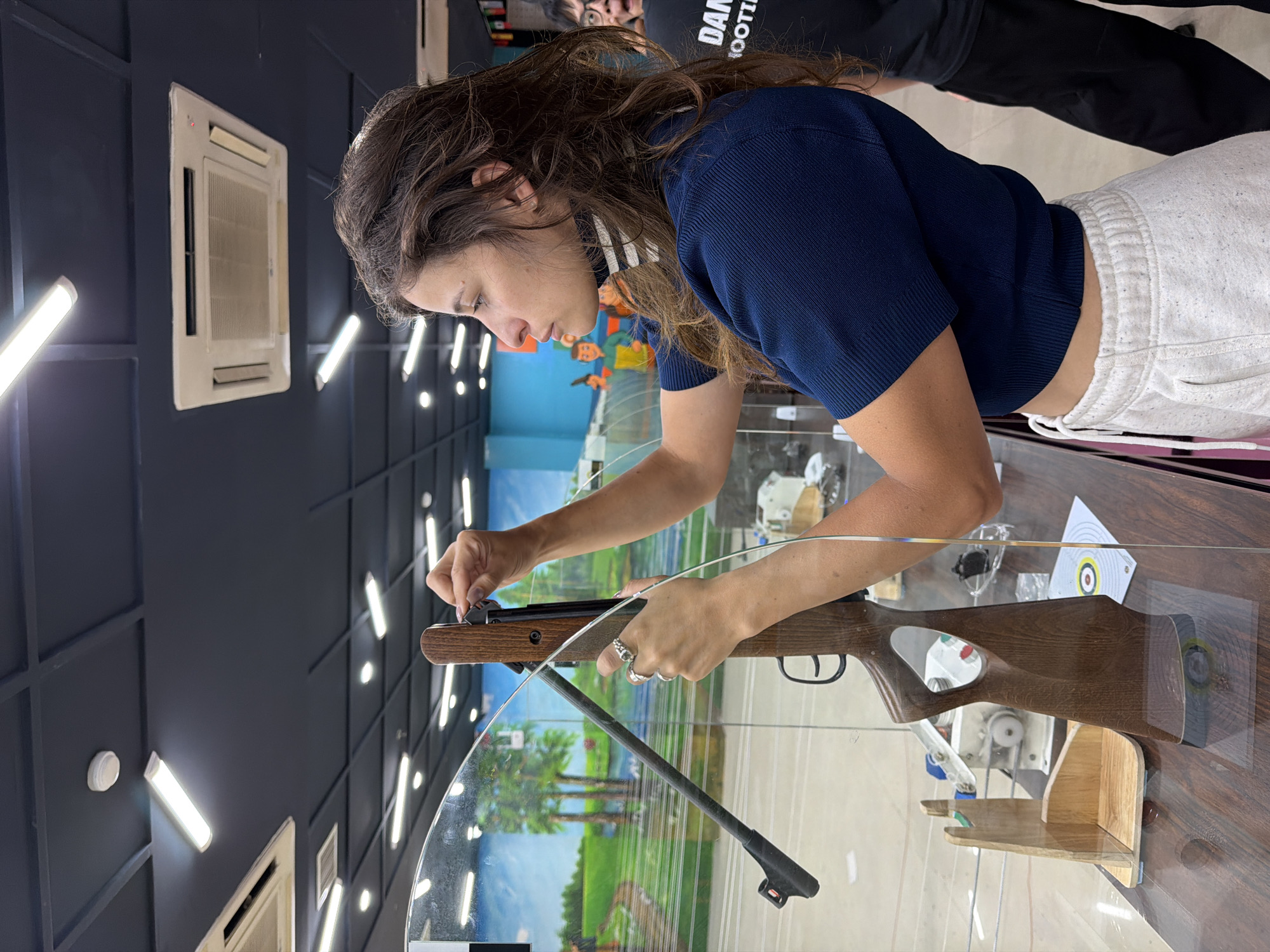 The image depicts a woman in a dark blue cropped sweater and light-colored sweatpants adjusting a rifle on a wooden table. The rifle, made of wood and metal, is supported by a clear plexiglass barrier. Behind the woman, a man in a black t-shirt with "DANI SHOOTING" printed on it is visible. The environment appears to be an indoor shooting range, with fluorescent lighting and what looks like a painted mural of a landscape on the back wall. The ceiling features a grid pattern and air conditioning units. The scene is set in the city of Đà Nẵng, Vietnam.