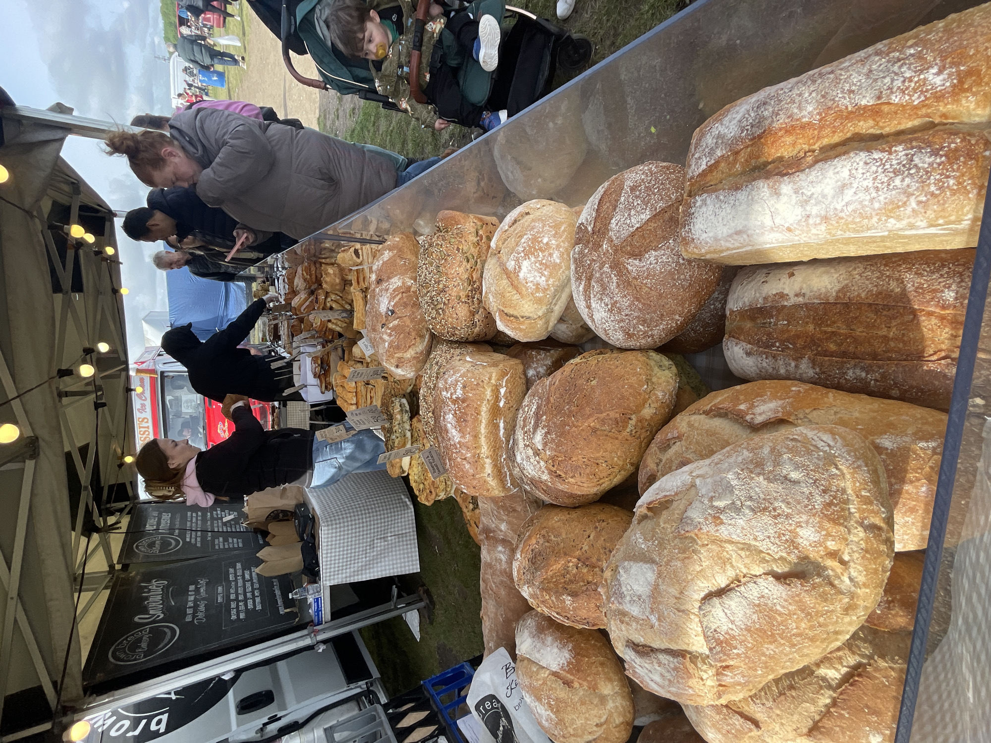 The image depicts a bread stall at a market in Sevenoaks, United Kingdom. A variety of loaves are displayed in front of a clear screen. Customers are examining the bread. A vendor stands behind the stall. A child in a stroller and other people are visible in the background. The market stall is covered. Other stalls and a van are also visible. The environment appears to be a field or open space.