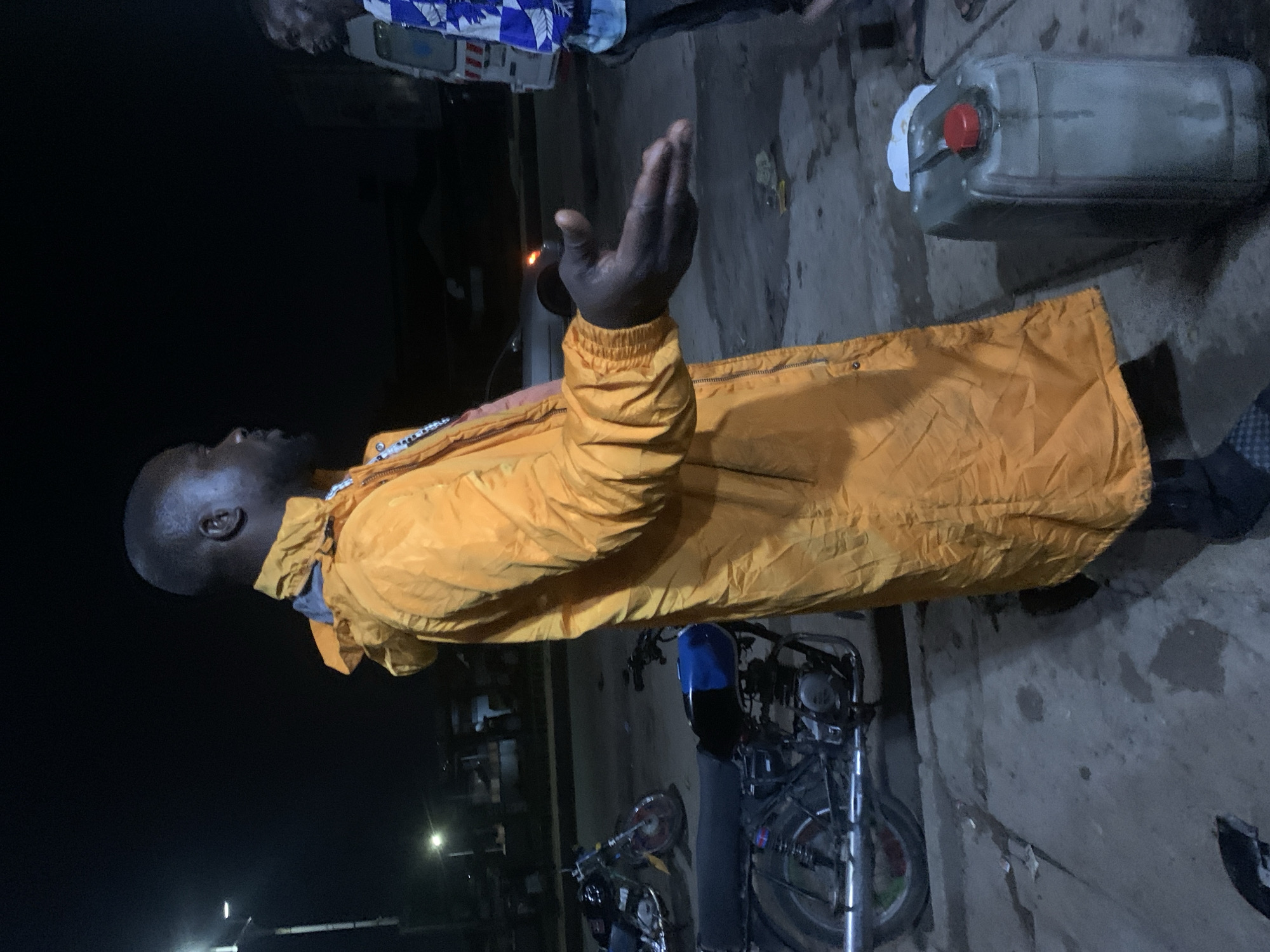The image, taken at night in Gura Top, Nigeria, features a man in a bright orange, possibly reflective, jacket standing outdoors. His right arm is raised, with his hand open and palm facing upwards, in a gesture that could signify prayer, supplication, or communication. His head is slightly tilted back, and he appears to be looking upwards or towards an unseen entity. The man has dark skin and a bald or shaved head.
Partially visible in the upper left background is another individual, wearing a blue and white patterned shirt, observing the scene. On the ground to the right, a grey plastic jerrycan with a red cap is placed, showing some white residue or spillage around its opening. A motorcycle is parked behind the central figure, with its handlebars and some mechanical components visible. The ground is a rough concrete or asphalt surface. The setting is dark, illuminated by ambient artificial lights in the distance, confirming it is nighttime. No distinct text is visible on any object.