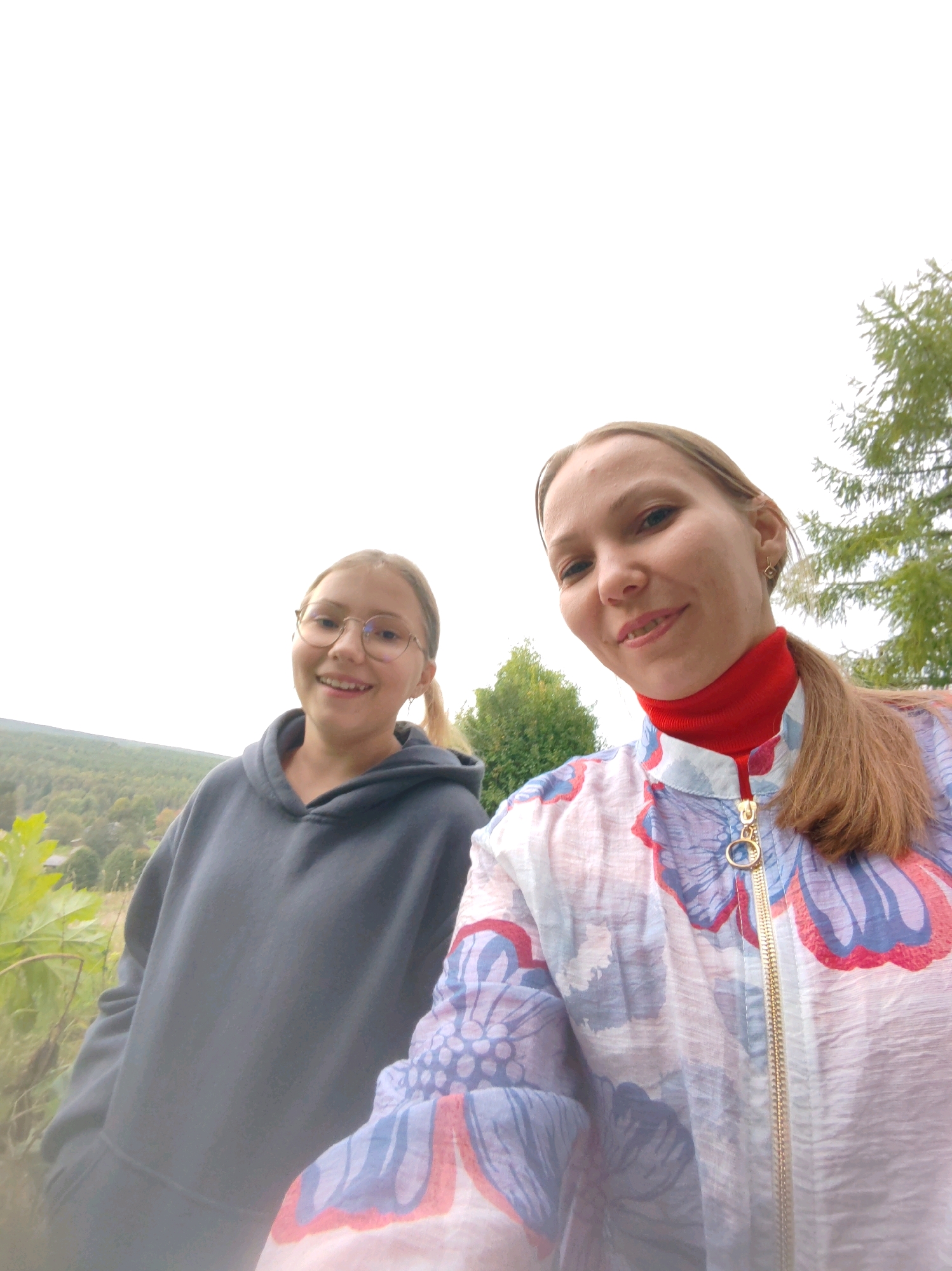 Two individuals, an adult female and a younger female, are captured in a close-up selfie outdoors. The adult, on the right, has light brown hair pulled back into a ponytail and is wearing a red turtleneck under a white jacket with a floral pattern in blue and red. She is smiling at the camera. The younger female, on the left, has blonde hair, wears glasses, and is dressed in a dark grey hooded sweatshirt. She is also smiling. They are positioned in front of a backdrop of trees and a hillside, suggesting a rural or natural setting. The sky is overcast. The location is not specified as the city of Чухлэм, Russia, but the background suggests a temperate climate.