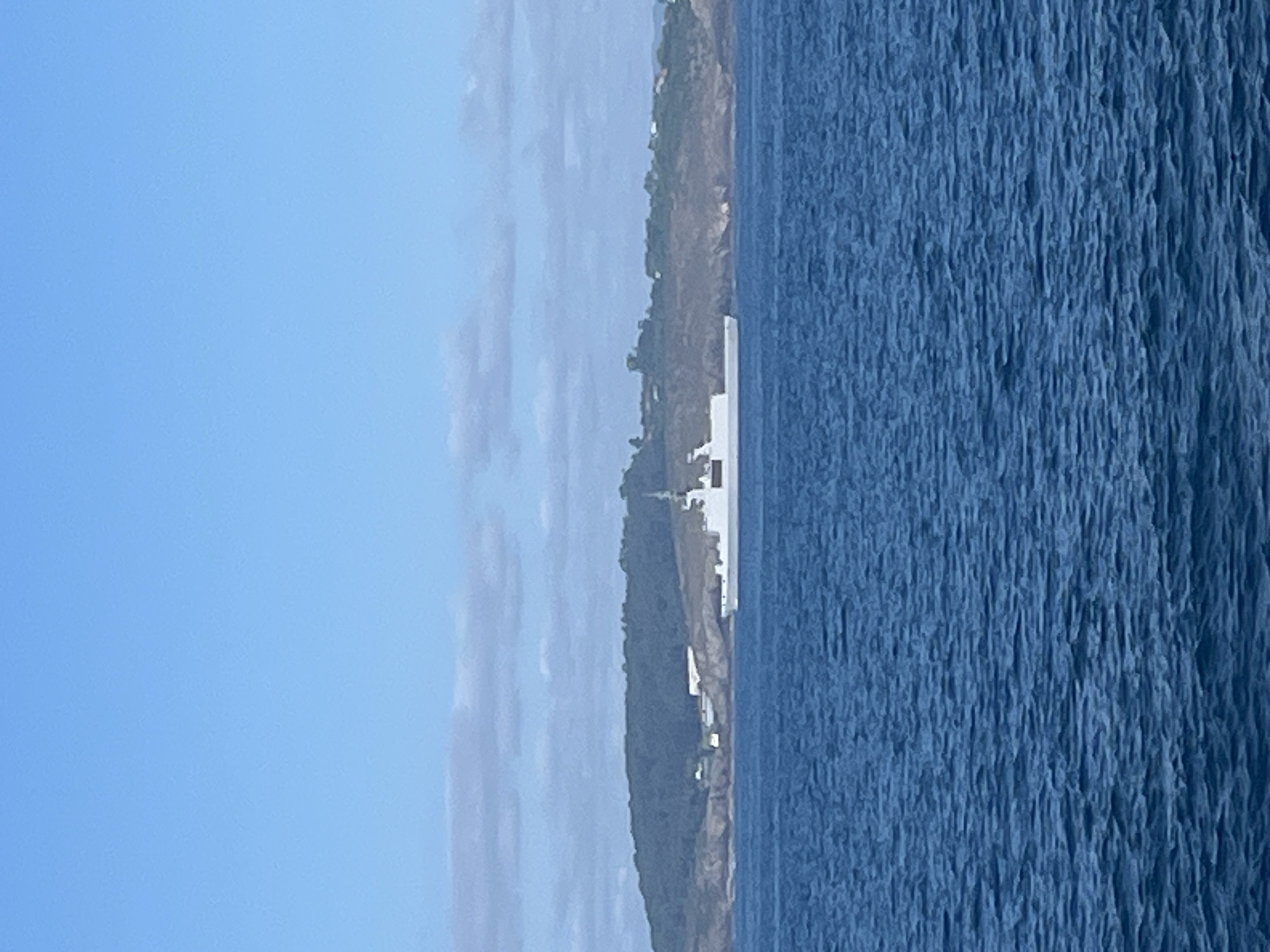 A white military ship is visible in the foreground, sailing in the blue ocean. In the background, a green, rocky island with some buildings on it can be seen. The sky above is a clear, light blue with some scattered clouds. The overall scene depicts a calm day at sea near the coast.