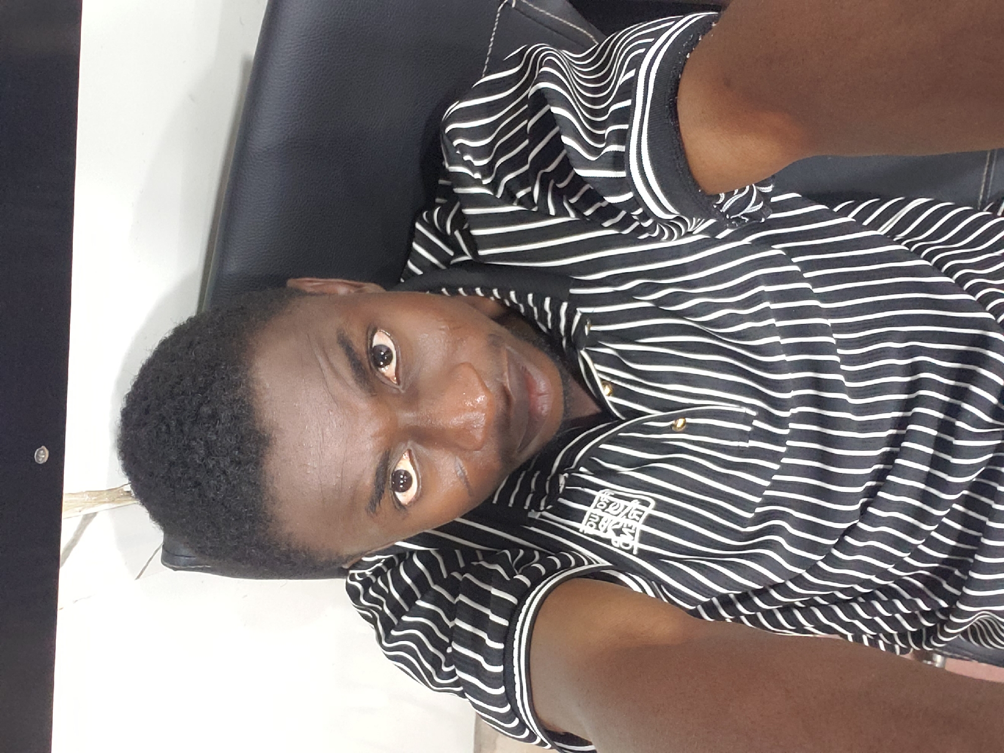 A dark-skinned man is featured in a selfie, looking directly into the camera with an open gaze and a neutral expression. He is seated indoors, likely in Adankolo, Nigeria, with his head slightly tilted. He wears a short-sleeved shirt with prominent black and white horizontal stripes, which ripple and curve due to the fabric's movement. The shirt has a dark collar and matching trim on the sleeves.
The man has short, dark, curly hair and some facial stubble or a short beard. He appears to be seated in a black upholstered chair or seat, which is visible behind and to the side of him. A plain white surface, possibly a wall, forms the background.
A visible detail on his shirt is a white rectangular patch on the left side of his chest. This patch contains stylized black text stacked vertically, which appears to spell "BELLO." The image is well-lit, suggesting it was taken during the day.