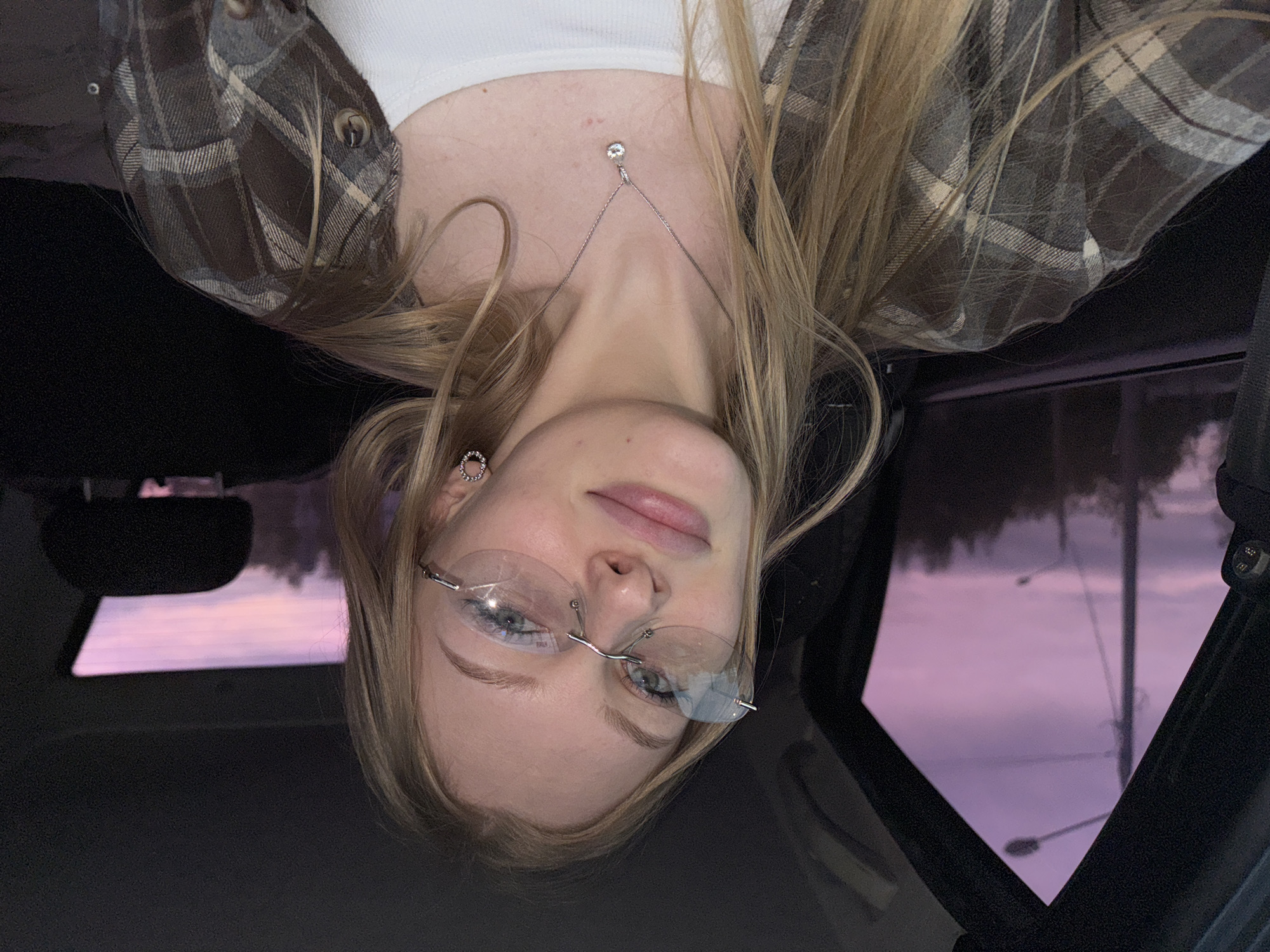 A young, fair-skinned woman with blonde hair is seen from an upside-down perspective, presumably taken from inside a vehicle. She is wearing rimless glasses, a white top, and a brown and tan plaid shirt. She is also wearing a necklace with a small diamond and circular earrings. The background visible through the car windows shows a purple and pink sky suggestive of dusk or dawn, with indistinct trees and streetlights visible. The location is suggested as Perm, Russia, but without further context, this is not verifiable in the image. The interior of the car is dark, with parts of the dashboard, steering wheel, and window frame visible.