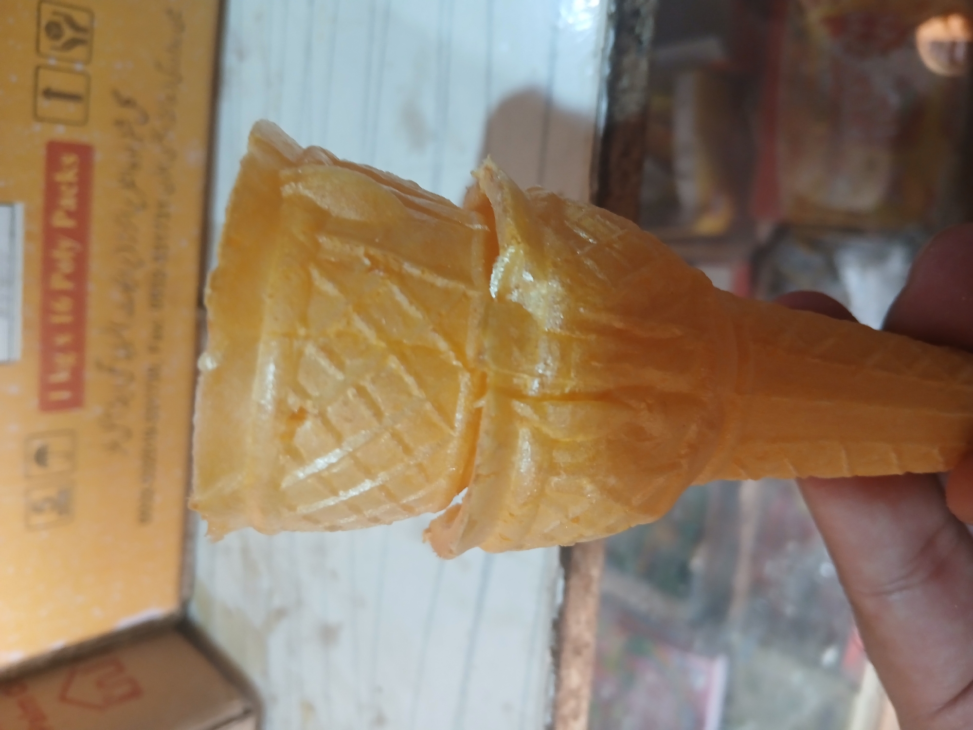 A close-up, slightly angled shot captures two empty, golden-brown ice cream cones stacked on top of each other and held in a person's hand. The cones have a textured, waffle-like pattern. In the background, out of focus, is a yellow box with some text and symbols, possibly indicating product information. Below the box, a blurry shelf or counter with various items is visible, and a reflective surface suggests a glass or plastic display case. The overall environment appears to be a shop or market, with the text on the box suggesting a location in Peshawar City Tehsil, Pakistan.