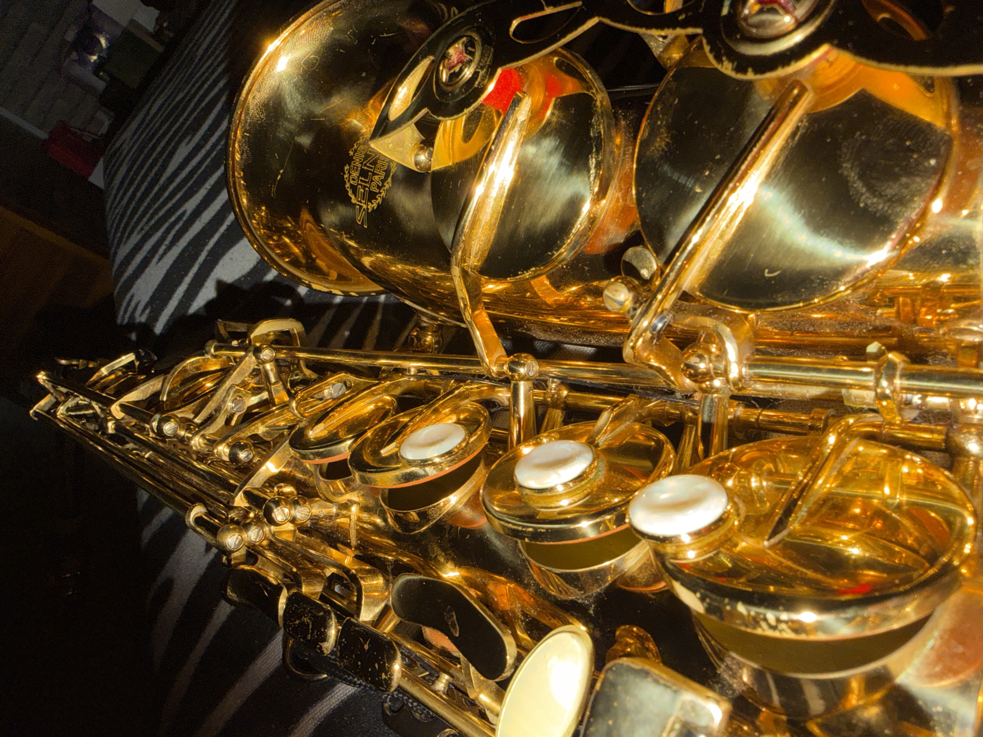 A close-up, high-angle photograph showcases the golden brass body of a saxophone, resting on a black and white striped surface, which appears to be a fabric cover. The instrument's intricate key mechanisms, including numerous levers, rods, and circular pads with white, pearl-like tops, are prominently featured. The highly polished brass reflects bright, artificial light sources, creating strong highlights and deep shadows that emphasize its metallic sheen and contours.
Visible text etched on the bell of the saxophone clearly identifies it as a "DENIS SELIN PARIS" instrument. Some minor dust or smudges are visible on the brass surfaces and around the key pads, adding to the realistic detail. The overall composition focuses tightly on the instrument, with the background elements slightly out of focus. The scene depicts a stationary musical instrument indoors, likely in Velingrad, Bulgaria.