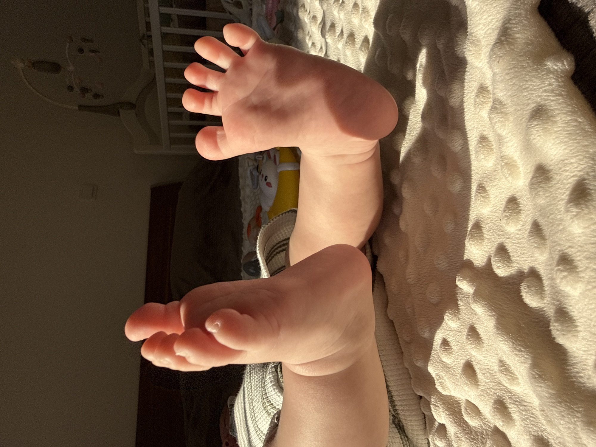 A close-up, high-angle shot shows the feet of a baby wearing striped bottoms. One foot is positioned on a textured cream-colored blanket, with the toes pointing upwards. The other foot is raised and slightly angled towards the left side of the frame. To the left of the feet, a baby crib and a wall are visible. The scene is lit by natural light, creating strong shadows on the blanket and the baby's skin. There is no indication that this image was taken in the city of Bar, Montenegro, or in any specific location.