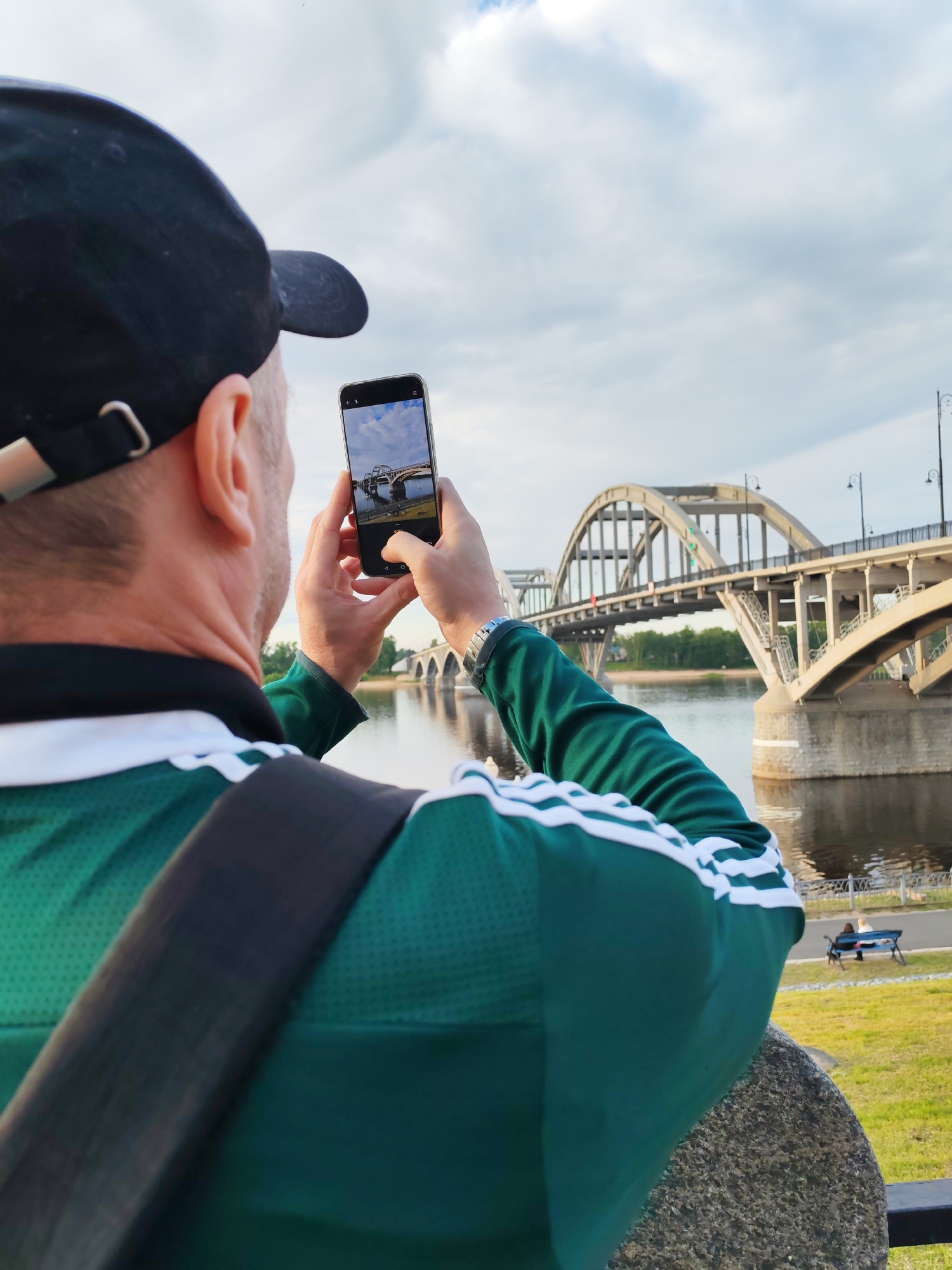 The image shows a man taking a picture of a bridge with his smartphone. The man is wearing a green shirt with white stripes on the sleeves and a black baseball cap. He is holding the phone in his right hand, and his left hand is supporting the phone. The phone's screen displays a picture of the bridge, indicating that he is framing his shot.
The bridge is a large concrete structure with several arched spans. It spans a wide river, and trees can be seen on the opposite bank. The sky is cloudy, suggesting it might be late afternoon or early morning. The overall scene is calm, with a sense of peacefulness.
In the background, there are a few people sitting on a bench by the river, enjoying the view. The lighting suggests it is daytime. The presence of a river, a bridge, and a park-like setting points to a public space, possibly a scenic spot for tourists or locals.
No visible text is present in the image. The man's focus is on capturing the beauty of the bridge and its surroundings. The image conveys a sense of appreciation for the scenery and the act of memory-making through photography. The location context "Bol'shie Mkhi, Russia" suggests a specific geographical setting for this scene.