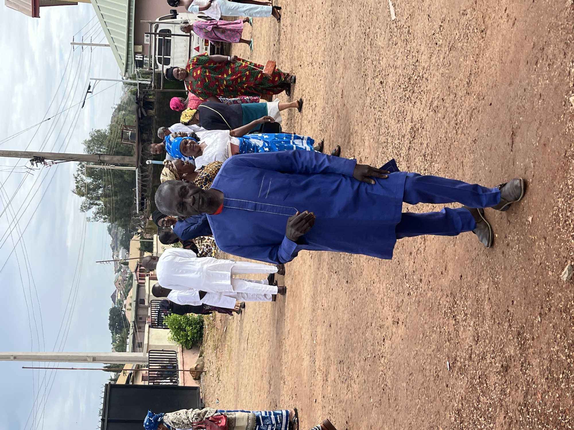 The image depicts an outdoor scene in what appears to be a residential area in Jos, Nigeria. The ground is covered with reddish-brown dirt and gravel. Several people are present, dressed in various traditional and modern attire.
In the foreground, a man in a royal blue tunic and trousers stands facing the camera. He has dark skin and is looking directly at the lens. His left hand is holding a small, dark object. His shoes are dark brown loafers.
Behind him, a group of people are gathered. To his left, several women are visible, some wearing headwraps and colorful patterned dresses. One woman in a white top and blue patterned skirt is looking towards the man in blue. Further to the left, a woman in a patterned outfit and a blue headscarf carries a red handbag.
To the right of the man in blue, other individuals are seen. A woman in a vibrant red and green patterned dress stands prominently, holding a small brown object in her right hand. Behind her, a white truck with a partial logo is visible, suggesting a commercial or community setting. In the background, houses with varying architectural styles are scattered across a gentle slope, characteristic of Jos. Utility poles and power lines crisscross the sky. The overall impression is a community gathering or event taking place on a somewhat overcast day.