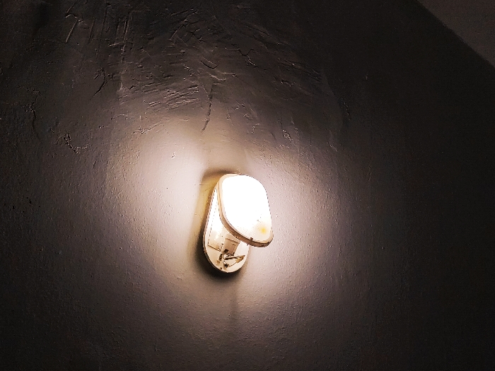 A wall-mounted light fixture illuminates a textured gray wall. The fixture is white with a rounded, rectangular shape. The light source is visible and bright, casting a circular glow around the fixture and onto the surrounding wall. The wall itself has a rough, uneven surface with visible trowel marks or a similar texture. The lower right corner of the image is dark, suggesting a corner or edge of a room.