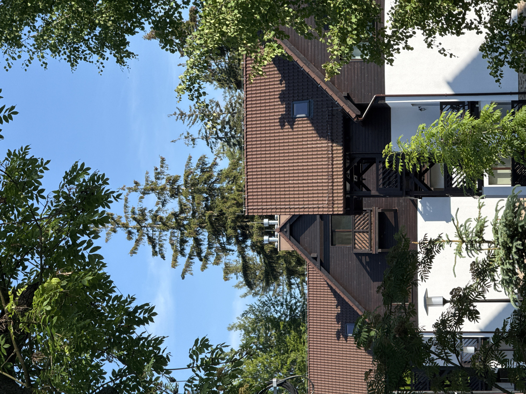 The image depicts the exterior of a multi-story building with a dark wooden facade and a brown tiled roof. The building has several balconies with wooden railings and dark framing. Two chimneys with silver caps are visible on the roof, along with a small skylight. In the foreground and framing the building are lush green trees with dense foliage. The sky is a clear, bright blue. In the lower part of the image, white walls are visible, and a modern-style lamp post with a white rectangular shade stands among the trees. The scene appears to be a residential or hotel building set within a natural, wooded environment. This is a view of the "Willa u Stasi" located in Szklarska Poręba, Poland.