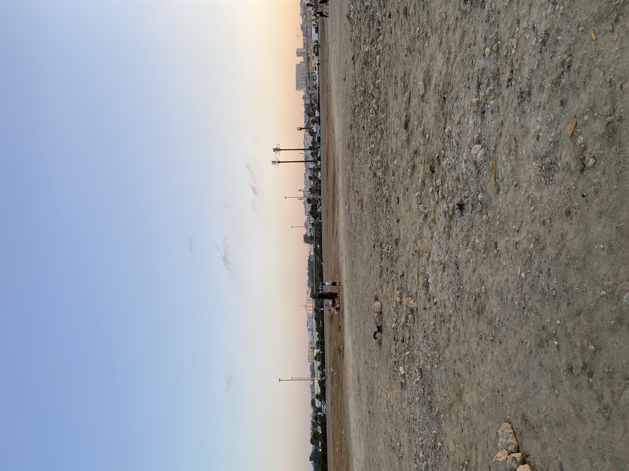 The image depicts an outdoor scene in Mohammedia, Algeria, under a clear sky with a gradient of blue and pale orange, indicative of either dawn or dusk. The foreground is a barren, rocky, and dusty terrain, with scattered pebbles and larger stones. A few pieces of litter, including a plastic bottle, are visible. In the mid-ground, a small group of people, including adults and children, are gathered. They appear to be engaged in leisure activities. Further in the background, a road runs horizontally, with vehicles parked and in motion. Beyond the road, a cityscape unfolds, characterized by buildings of varying heights and architectural styles, and palm trees. Utility poles and two distinctive tall structures resembling pylons are also present. The overall impression is of an urban periphery transitioning from undeveloped land to a populated city.
