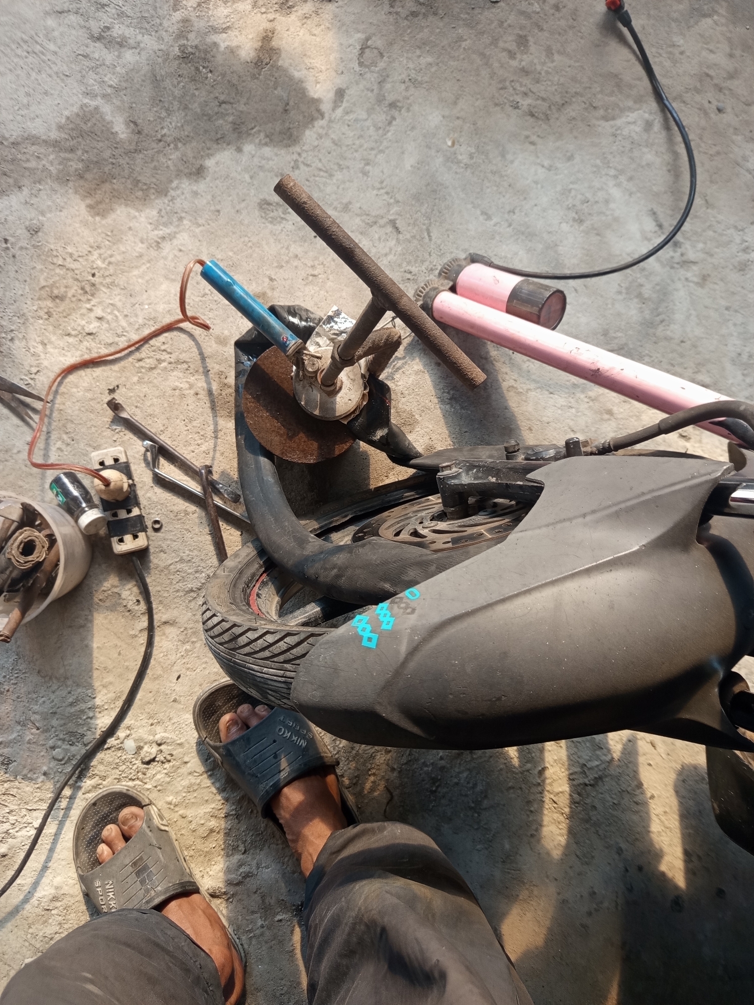 A high-angle, medium shot captures a person's feet wearing black sandals on a concrete surface in what appears to be a workshop or outdoor area in Ngablak, Indonesia. The person is wearing black trousers. In the foreground, the feet and lower legs are visible. The scene is cluttered with various objects, including a motorcycle wheel and part of its body, tools such as wrenches, a grinding disc, electrical extension cords plugged into a power strip, and some pink pipes. The concrete ground shows signs of wear and some water stains. The lighting suggests it is daytime, with shadows indicating the direction of the sun. The overall impression is one of a work-in-progress or a storage area for tools and equipment.