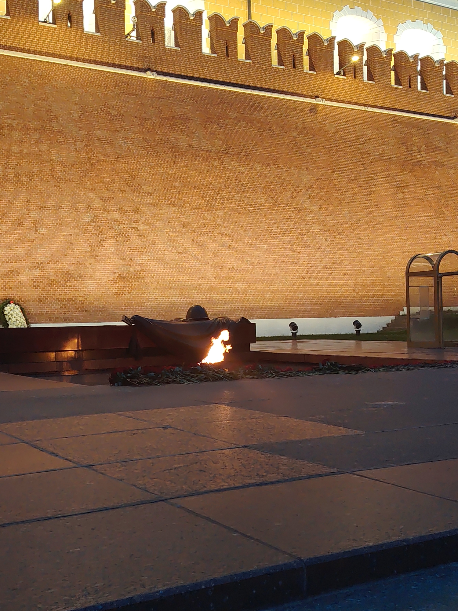 An outdoor nighttime scene features a memorial with an eternal flame. The memorial is situated in front of a large, textured brick wall, which is illuminated. A black tarp drapes over a portion of the memorial, and a helmet rests on top of it. The flame is visible and appears to be actively burning. Wreaths of flowers are placed around the base of the memorial and to the left. In the background, there are a few spotlights and a glass-enclosed structure. The ground in the foreground is paved with large square tiles. The overall impression is one of solemnity and remembrance.