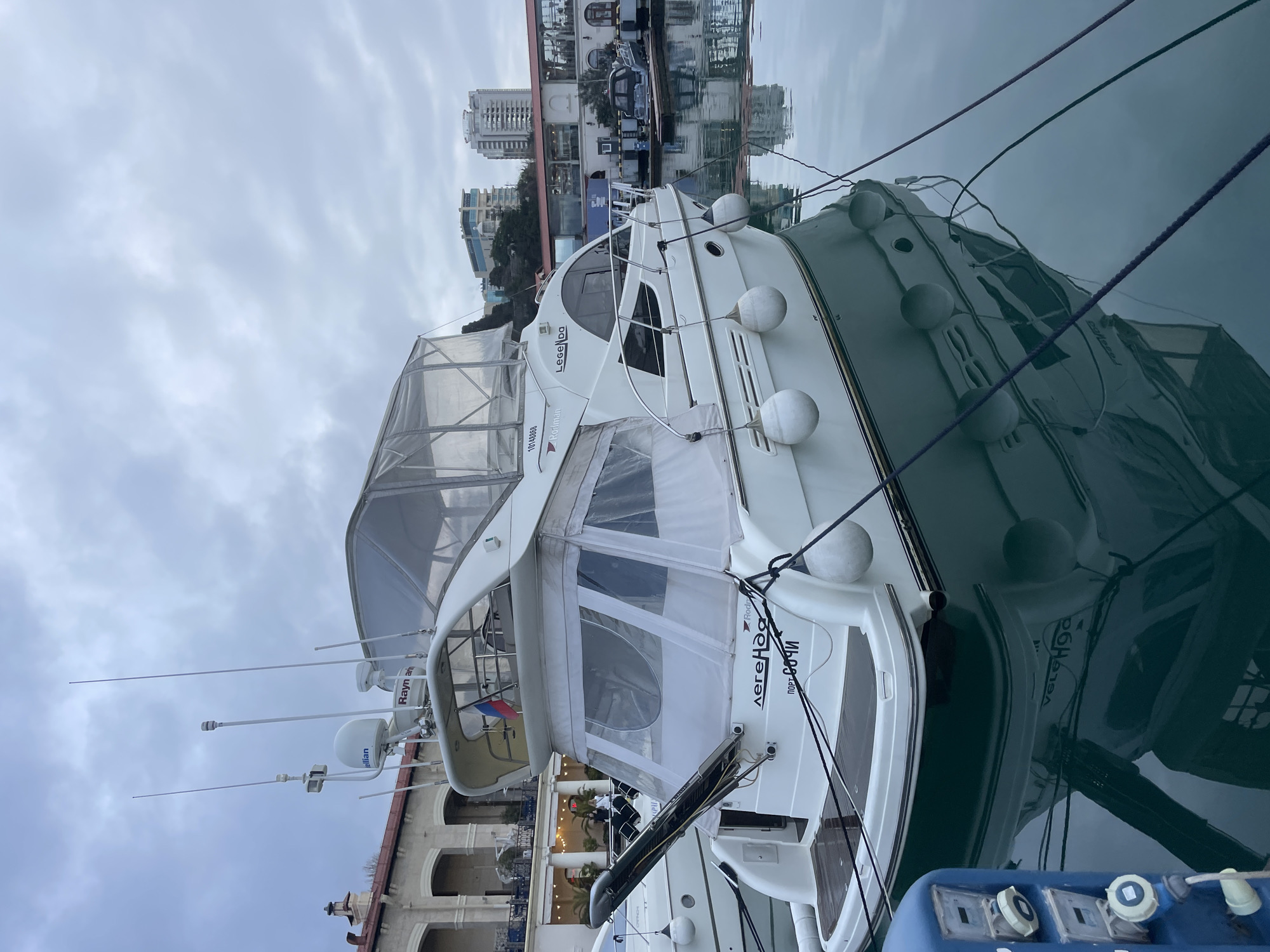 A white motor yacht named "Легенда" (Legenda) is moored in the Port of Sochi, Russia, under an overcast sky. The yacht's stern displays "Легенда" and "Порт Сочи" in Cyrillic, along with the hull number "10148858" and manufacturer "Roftman". A small Russian flag is mounted on the stern. The vessel is equipped with a tender lift/davit, multiple antennae, and two radar domes, one bearing "Kjellian" and the other "Raymarine" branding. A clear vinyl enclosure covers parts of the upper deck and stern. Six white cylindrical fenders hang along the starboard side. The yacht is secured to a blue mooring pedestal via black lines, and its reflection is visible in the calm, dark green water.
In the background, to the left, is a light-colored building with arched windows and balconies, characteristic of the Sochi Sea Port Terminal, with illuminated interiors and at least two indistinct figures visible on a balcony. To the right, beyond the pier, modern high-rise buildings with glass facades stand on a hillside. Another small, dark-colored boat is visible on a lift at a distant pier. The overall scene depicts a marina environment with urban architecture under a uniformly cloudy sky.