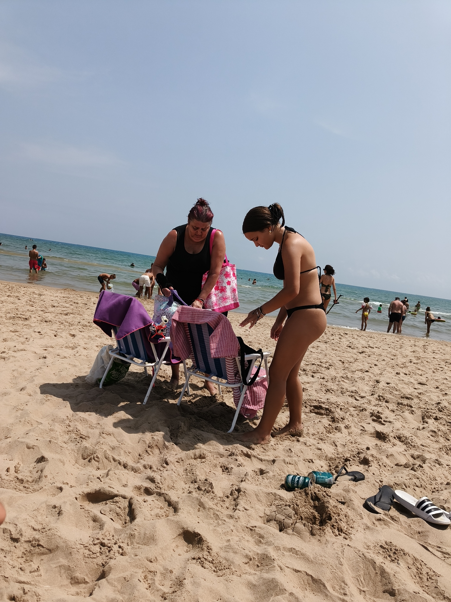 The image depicts a beach scene in Grau I Platja, Spain. Two women are in the foreground, with one in a black tank top and the other in a black bikini. They are standing near two folding chairs with towels draped over them. The woman in the bikini appears to be reaching for something on the chairs. In the background, several people are in the ocean, and the beach extends towards the water. The sand is a light brown color, and the sky is clear and blue. Several cans of what appear to be beer are on the sand near the chairs, along with a pair of flip-flops. The overall impression is a typical summer day at the beach.