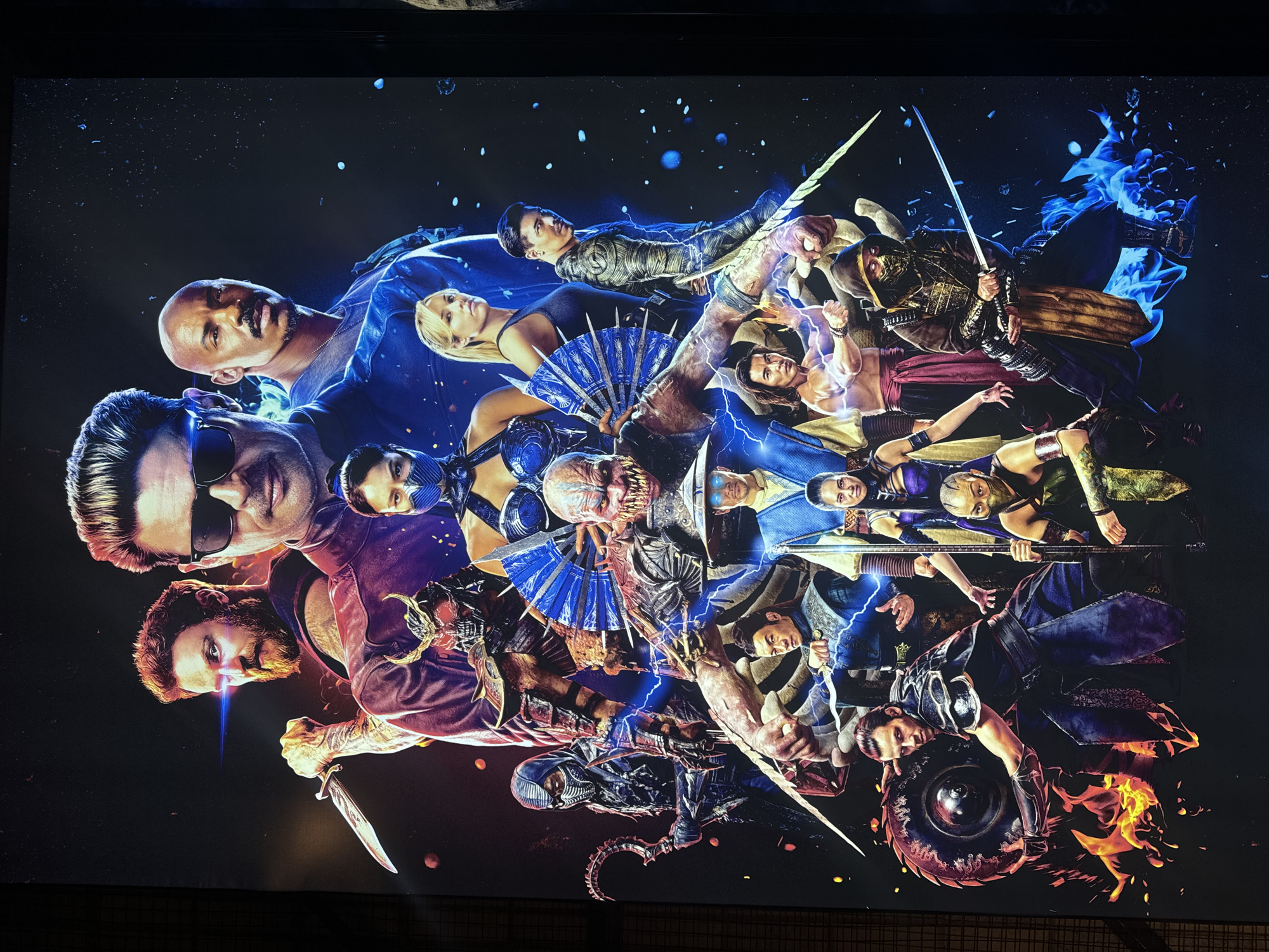 The image is a poster or print depicting characters from the video game franchise Mortal Kombat. Various fighters are arranged in a dynamic, overlapping composition. On the left side, a man with sunglasses and another with a beard and knife are prominent. In the center, there's a monstrous figure with blue fans. Several other characters with weapons and fighting stances are distributed throughout the image. Blue and red lighting effects and stylized imagery create a dramatic atmosphere. The background is dark with abstract elements suggesting cosmic effects. There is no evidence this image comes from Thu Duc City, Vietnam.