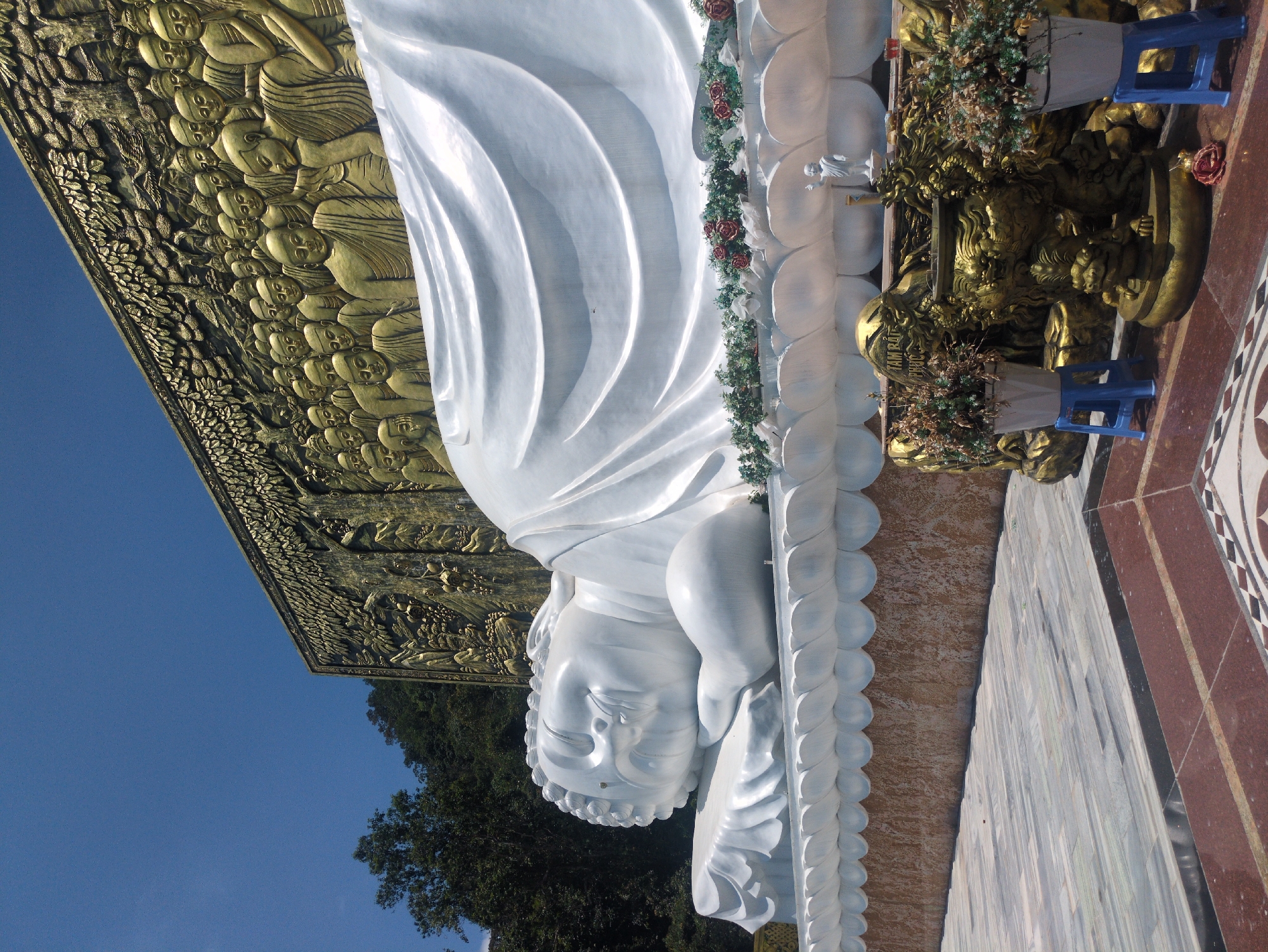 The image shows a large, reclining Buddha statue located on Phú Quốc Island in Vietnam. The statue is predominantly white, with intricate details on the head, body, and draped clothing. It is adorned with floral decorations. Above the statue is a golden bas-relief featuring many figures, likely depicting scenes from Buddhist scripture. The statue rests on a platform with marble-like tiling and ornate edges. There are smaller golden statues at the foot of the reclining Buddha, decorated with plants in pots set on small blue stools. The background reveals a clear blue sky and green trees, suggesting an outdoor setting.