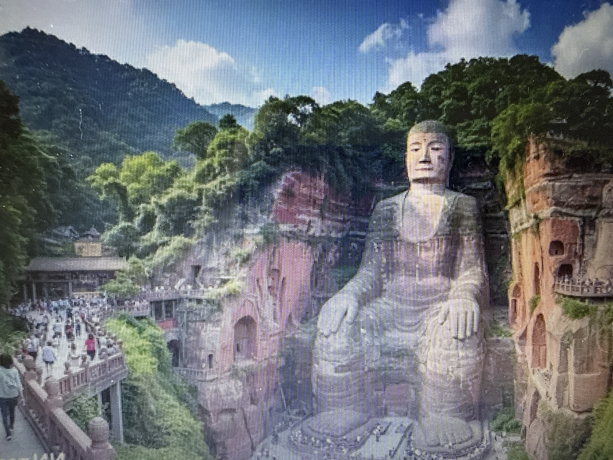 The media file shows a wide shot of a large, seated Buddha statue carved into a cliff face. The statue is made of stone and is weathered, with visible carvings and details. The Buddha is facing forward, with its hands resting in its lap. To the left of the statue, there is a busy tourist area with many people walking along paths and a bridge. There are also buildings and structures integrated into the cliff face. Green foliage covers parts of the cliffs and the surrounding landscape. In the background, lush green mountains rise up under a bright blue sky with scattered white clouds. The scene appears to be set during the daytime, likely a sunny day, given the brightness of the sky. The overall mood is one of awe and wonder, with the grandeur of the statue set against a beautiful natural landscape and the activity of visitors. No text is visible in the media file.